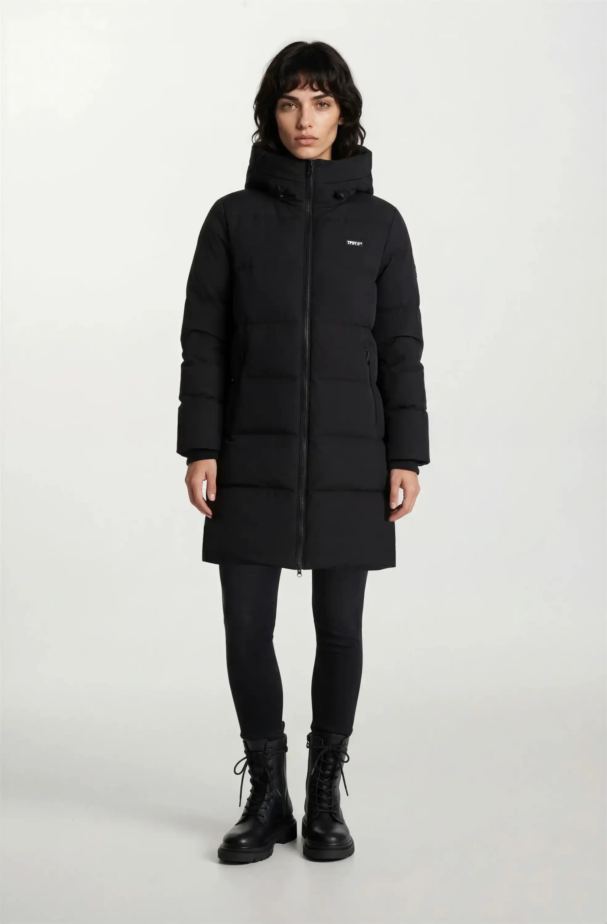 Eine Person steht mit dem Gesicht nach vorne und trägt einen trueprodigy Damen Parka mit fester Kapuze im Regular Fit, eine schwarze Hose und schwarze Schnürstiefel vor einem schlichten hellen Hintergrund.