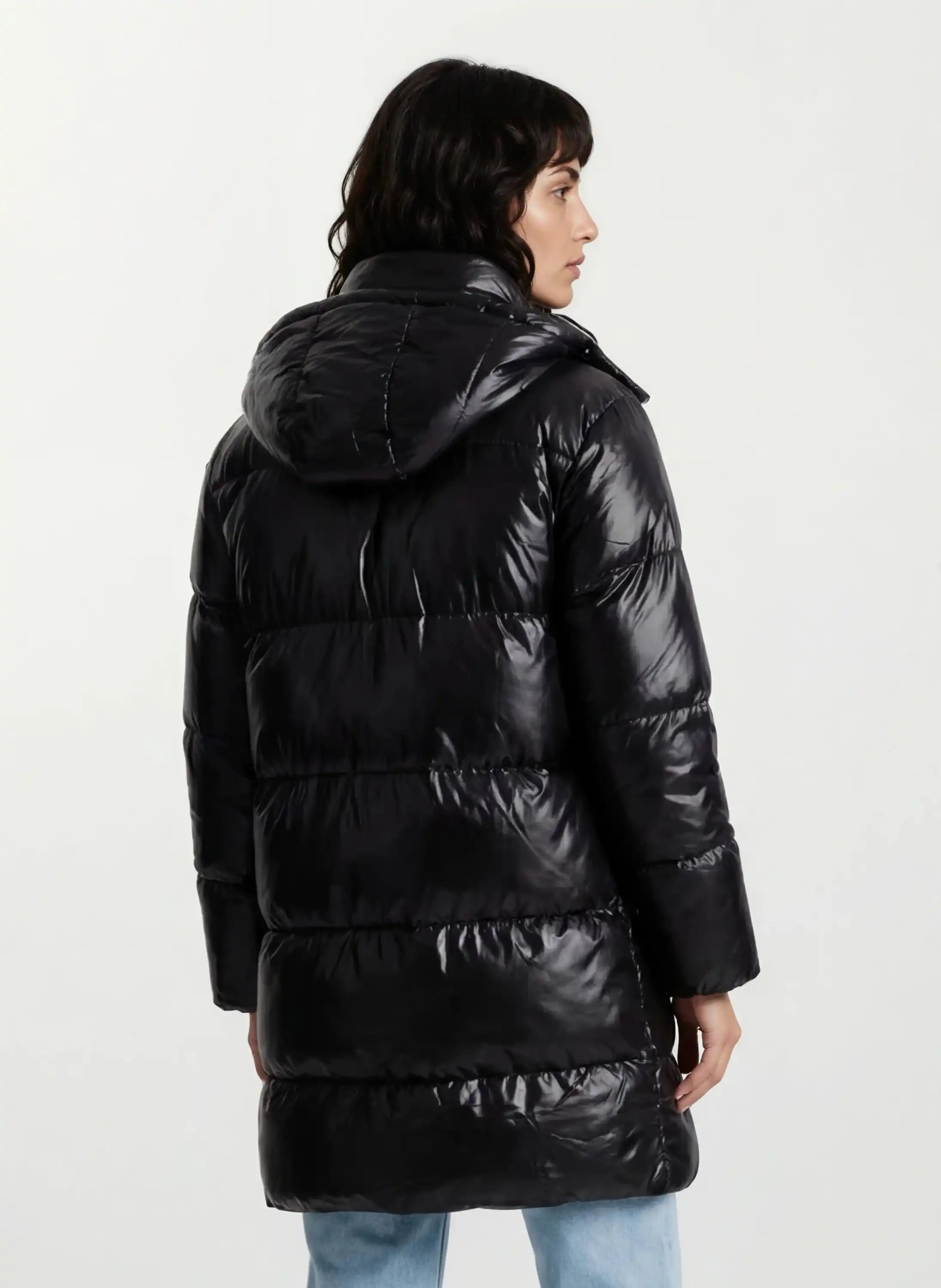 Eine Person mit dunklen Haaren ist von hinten zu sehen, die eine trueprodigy Damen Winterjacke mit hochglänzender Oberfläche im Regular Fit trägt. Der lange schwarze Mantel reicht bis zu den Oberschenkeln und ist mit einer hellblauen Jeans gepaart.