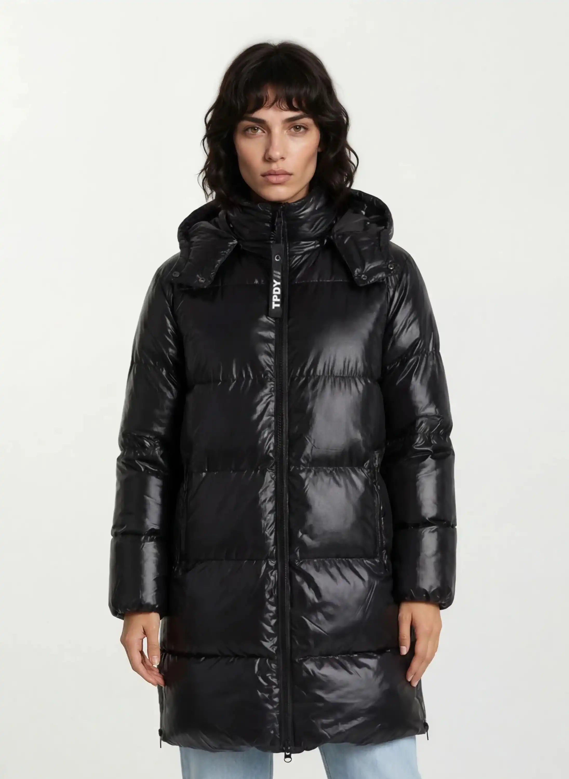 Eine Person, die die trueprodigy Damen Winterjacke mit hochglänzender Oberfläche im Regular Fit mit abnehmbarer Kapuze trägt, steht mit dem Gesicht nach vorne vor einem schlichten weißen Hintergrund. Der knielange Mantel hat einen Frontreißverschluss und eine glänzende Oberfläche.