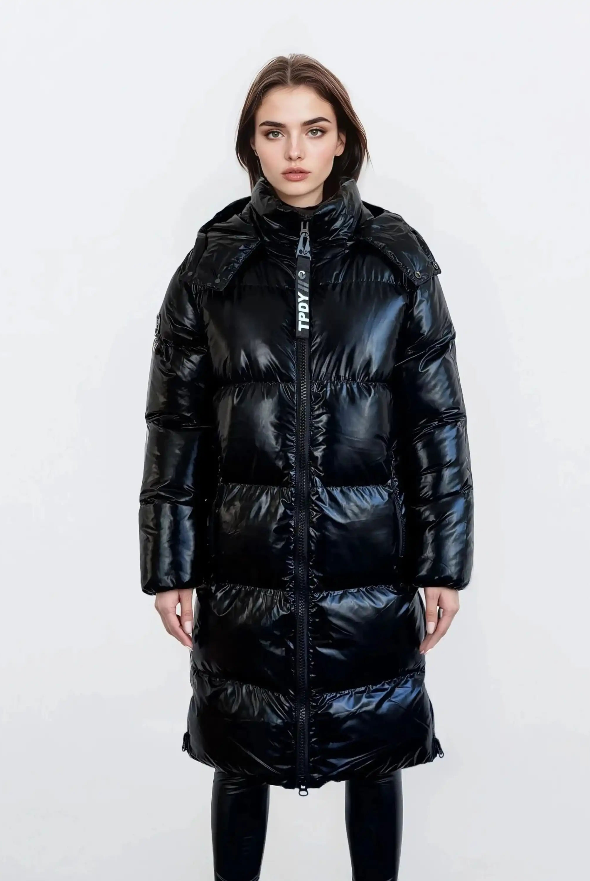 Eine Frau mit glattem braunem Haar und neutralem Gesichtsausdruck modelliert die trueprodigy Damen Winterjacke mit hochglänzender Oberfläche im Regular Fit, vorne mit Reißverschluss und abnehmbarer Kapuze, über einer schwarzen Hose vor einem schlichten weißen Hintergrund.