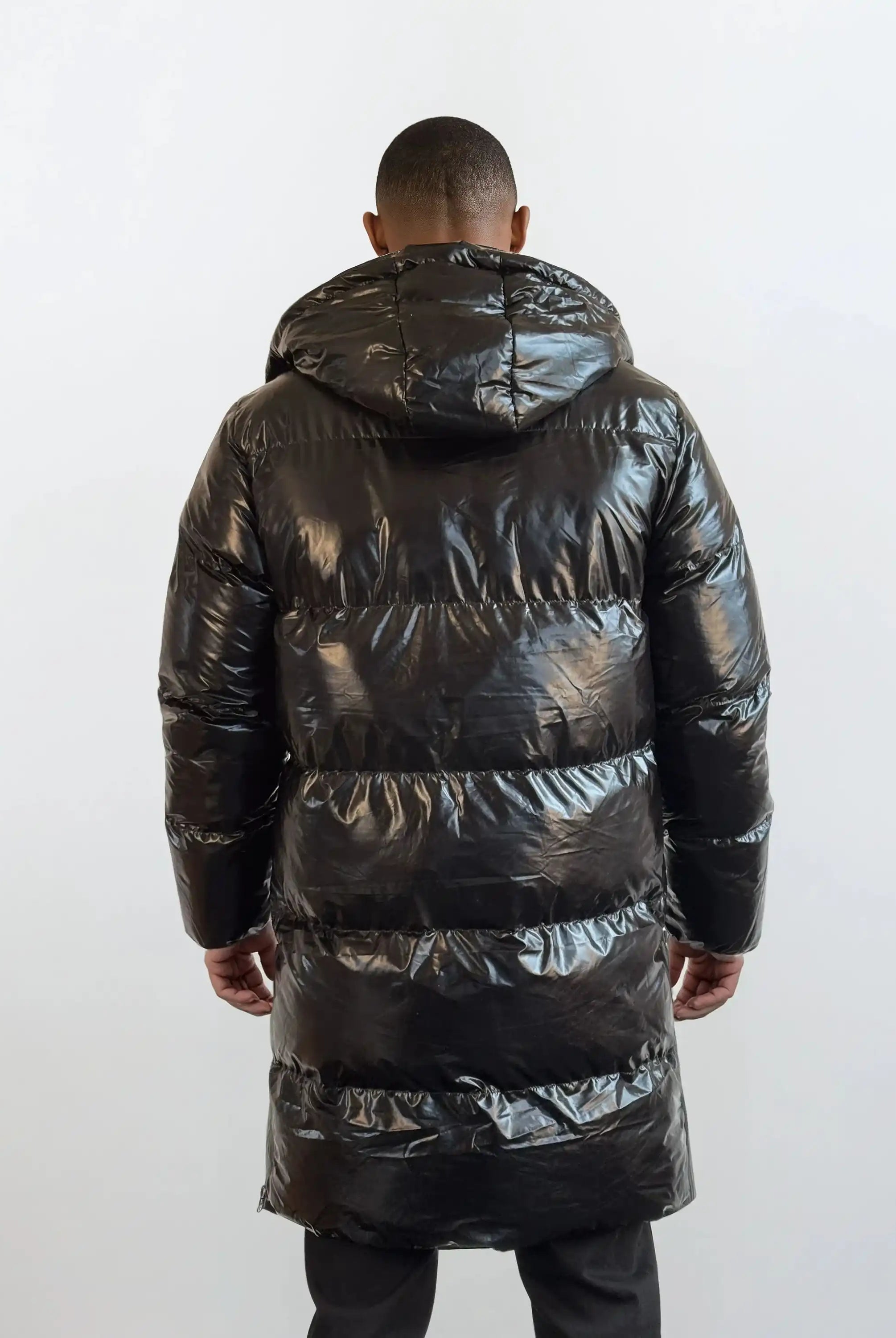 Eine Person blickt von der Kamera weg und trägt die trueprodigy Herren Winterjacke mit hochglänzender Oberfläche im Regular Fit. Die lange schwarze Pufferjacke mit Kapuze hebt sich von dem hellen, schlichten Hintergrund ab.