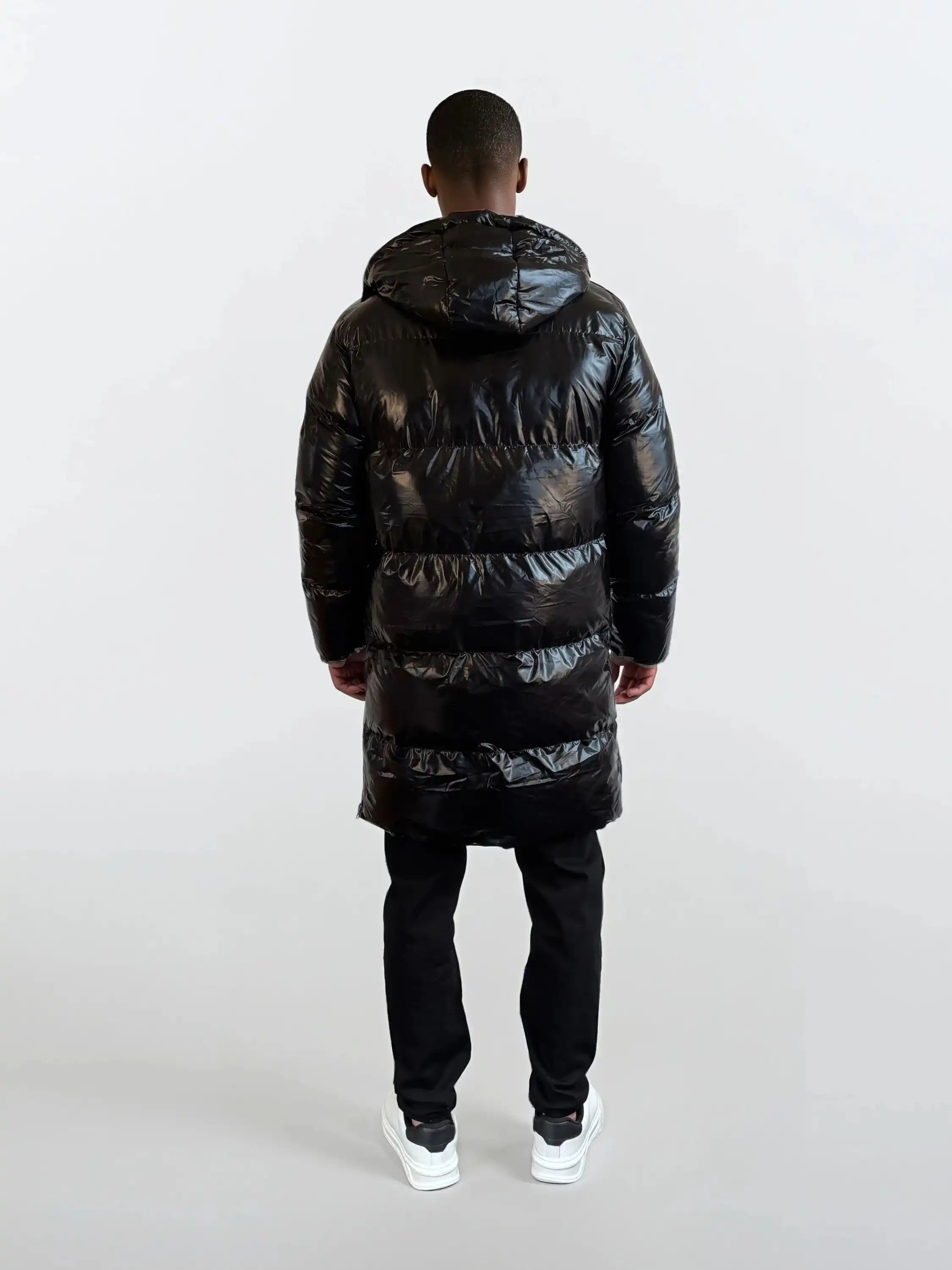 Eine Person steht von der Kamera abgewandt und trägt eine trueprodigy Herren Winterjacke mit hochglänzender Oberfläche im Regular Fit, eine schwarze Hose und weiße Turnschuhe. Der Hintergrund ist einfarbig und hell.
