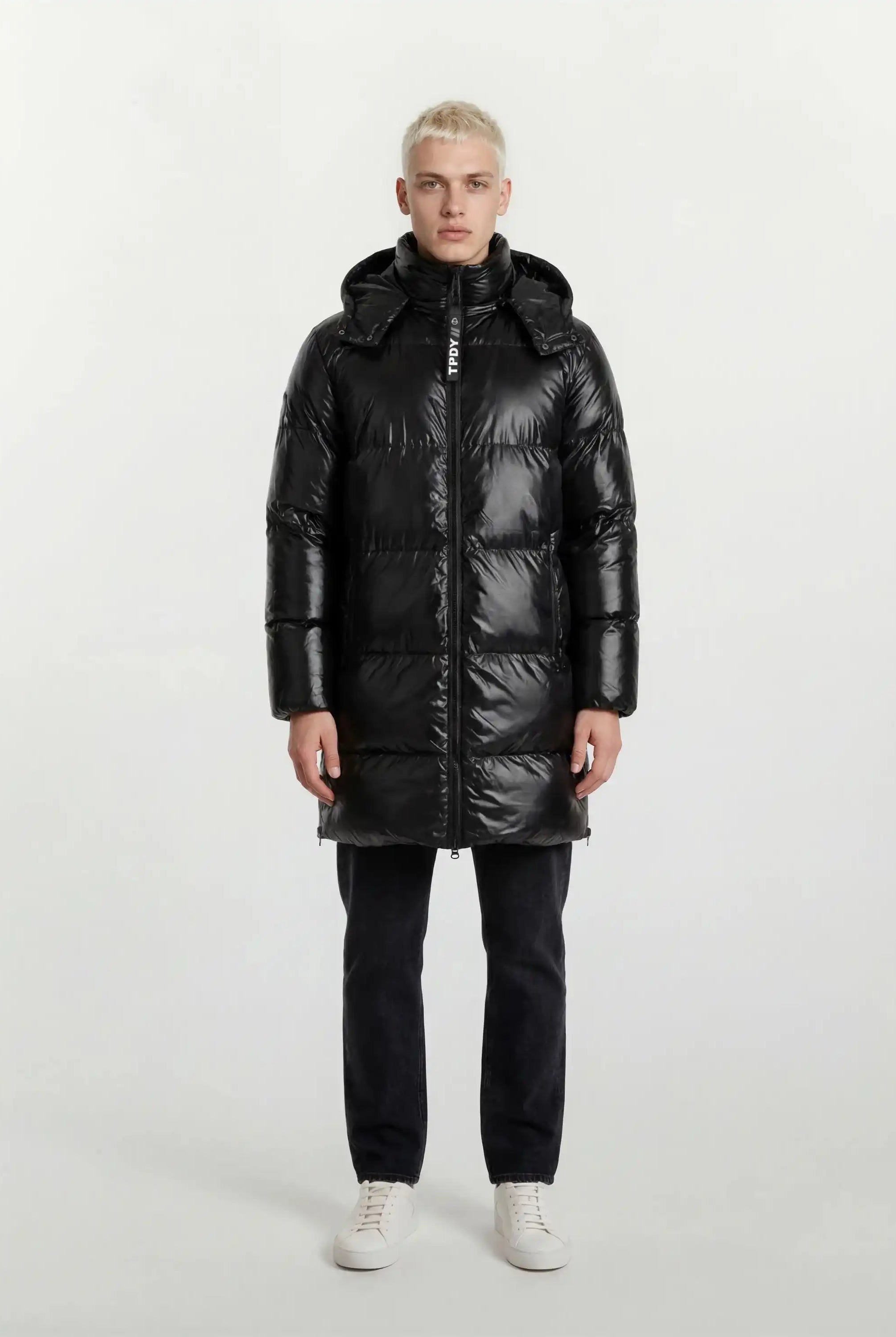 Eine Person steht mit dem Gesicht nach vorne und trägt eine trueprodigy Herren Winterjacke mit hochglänzender Oberfläche im Regular Fit, eine schwarze Hose und weiße Turnschuhe vor einem schlichten, hellen Hintergrund.