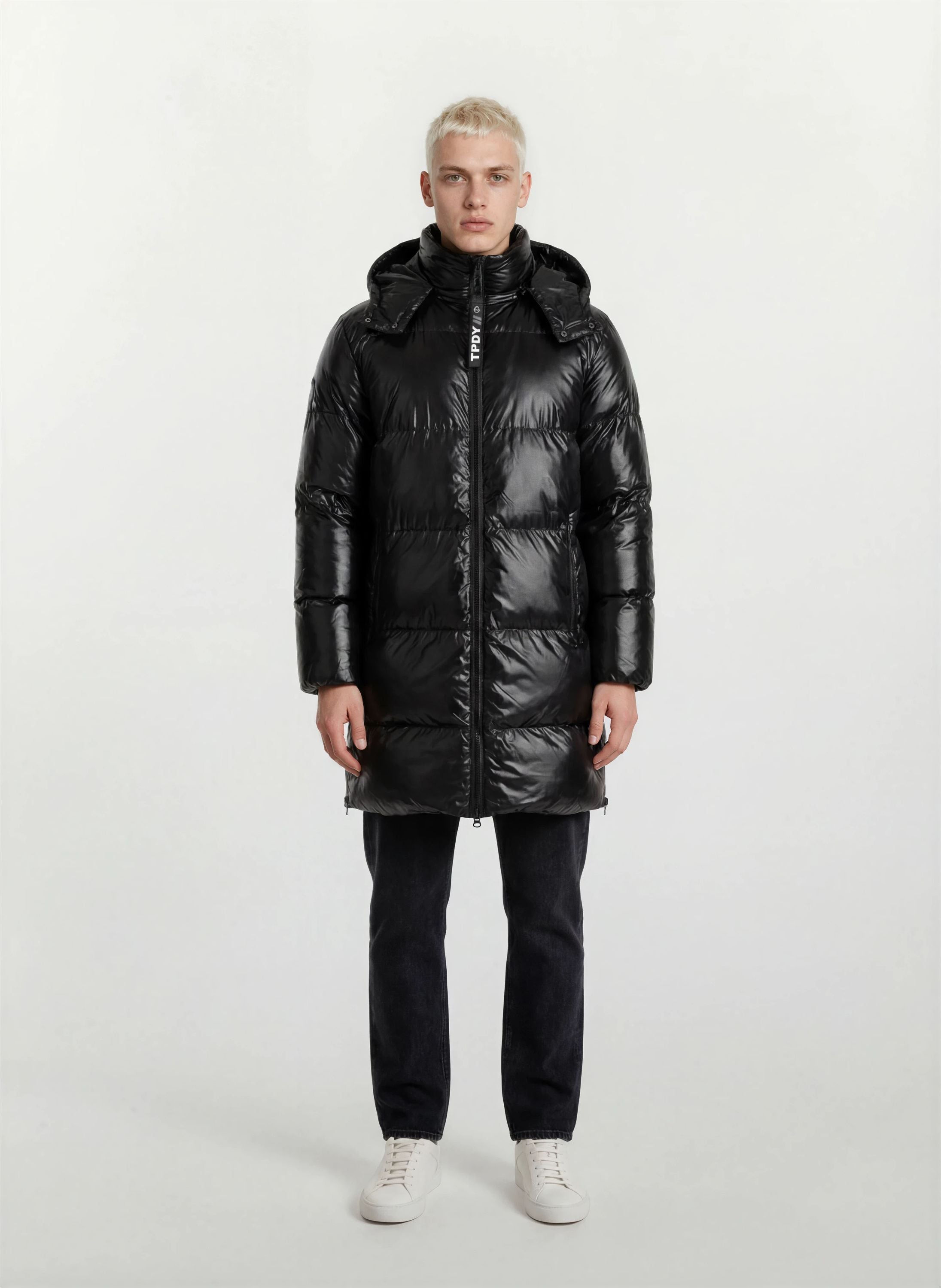 Eine Person steht mit dem Gesicht nach vorne und trägt eine trueprodigy Herren Winterjacke mit hochglänzender Oberfläche im Regular Fit, eine schwarze Hose und weiße Turnschuhe vor einem schlichten, hellen Hintergrund.