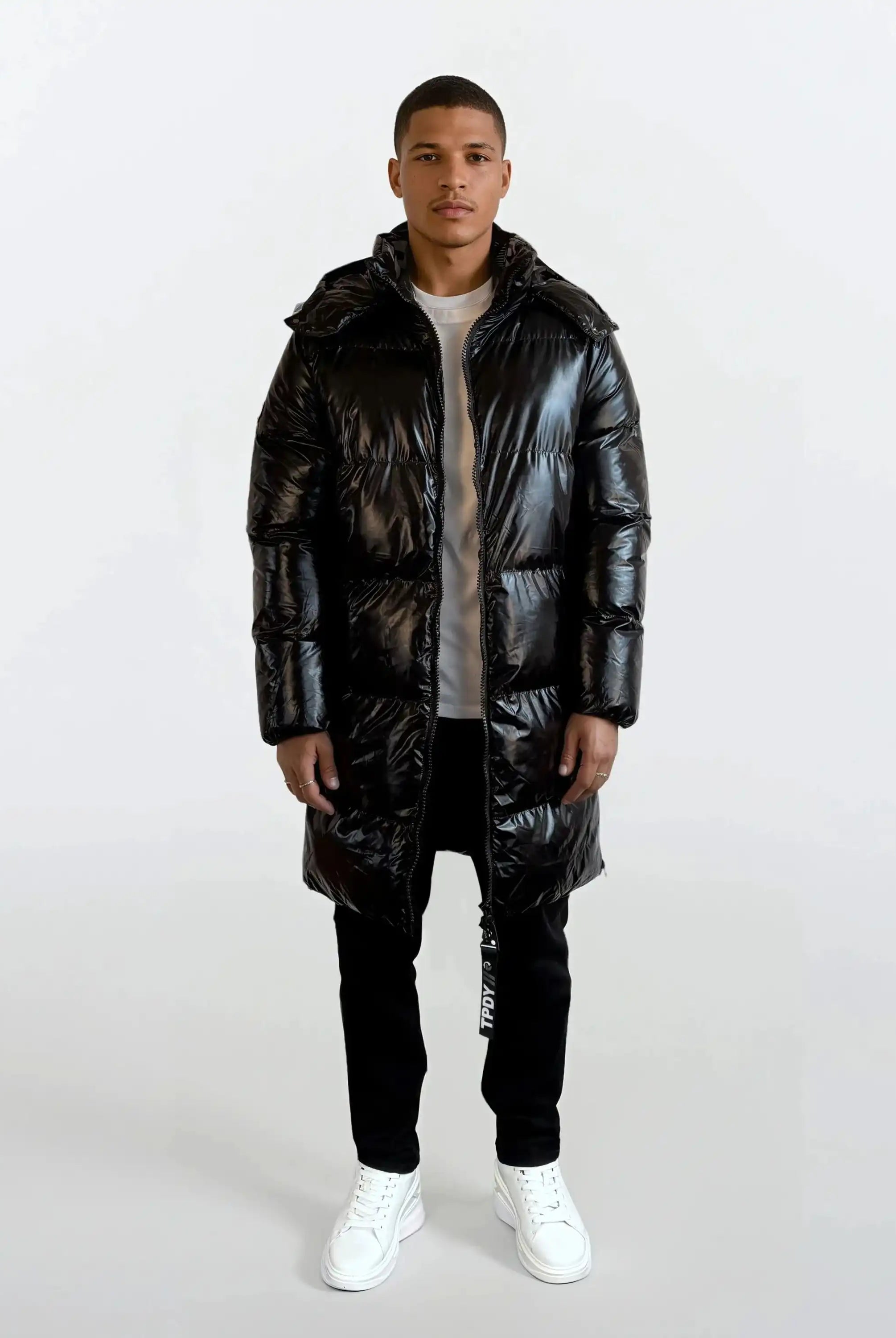 Eine Person steht mit Blick nach vorne in einer glänzenden schwarzen trueprodigy Herren Winterjacke mit hochglänzender Oberfläche im Regular Fit mit abnehmbarer Kapuze, schwarzer Hose, hellem Hemd und weißen Turnschuhen vor einem schlichten weißen Hintergrund.