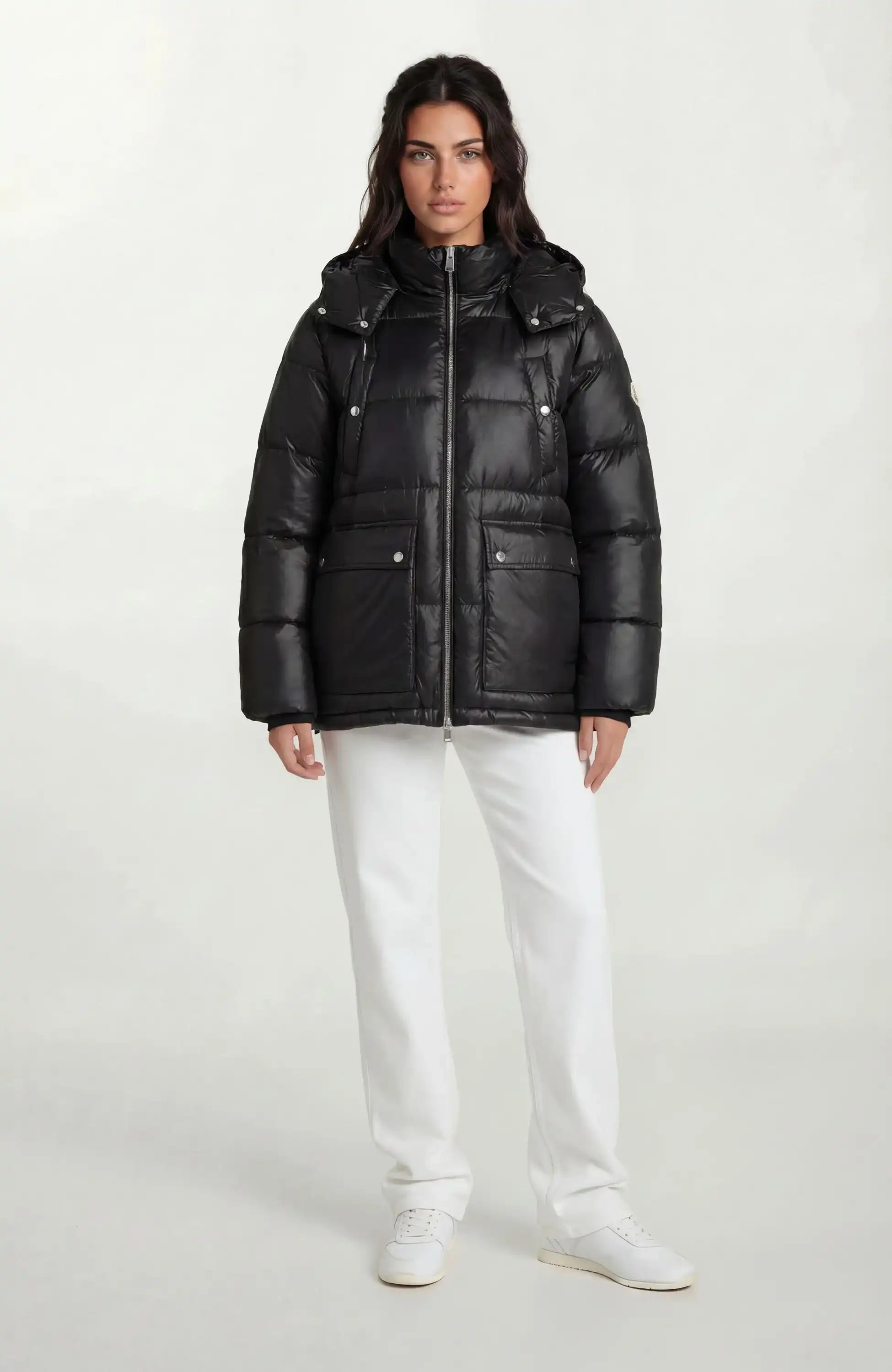 Eine Person steht mit dem Gesicht nach vorne und trägt eine trueprodigy Damen Winterjacke mit hochglänzender Oberfläche im Regular Fit, eine weiße Hose und weiße Turnschuhe vor einem hellgrauen Hintergrund.