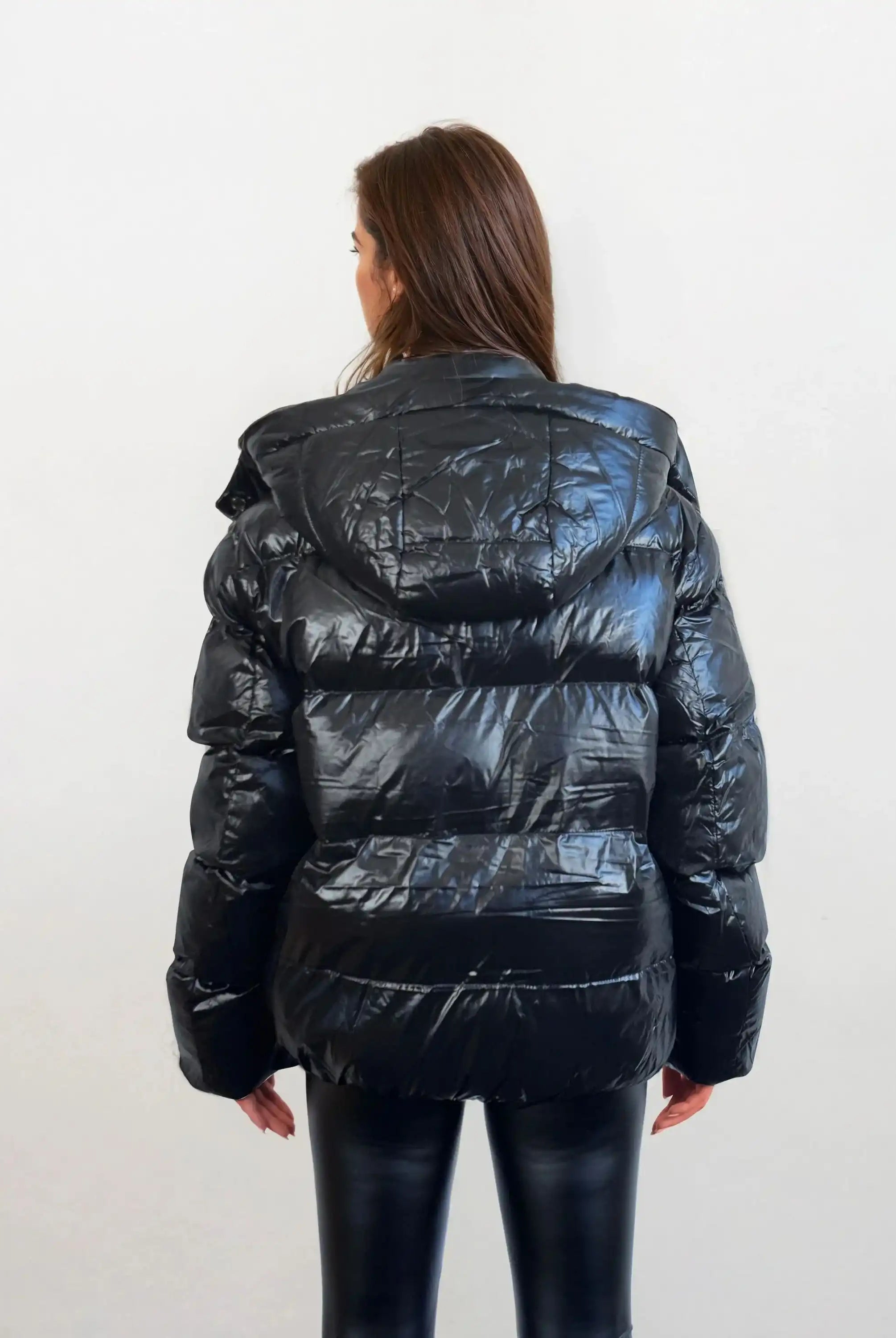 Eine Person mit langen braunen Haaren trägt eine trueprodigy Damen Winterjacke mit hochglänzender Oberfläche im Regular Fit und eine schwarze Hose, steht vor einem schlichten hellen Hintergrund und schaut von der Kamera weg.
