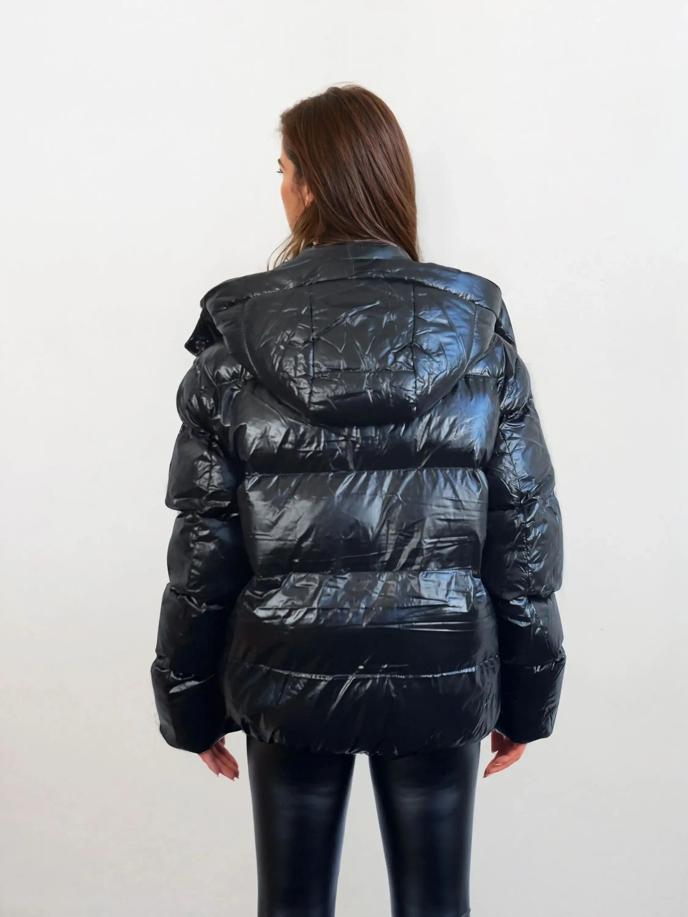 Eine Person mit langen braunen Haaren trägt eine trueprodigy Damen Winterjacke mit hochglänzender Oberfläche im Regular Fit und eine schwarze Hose, steht vor einem schlichten hellen Hintergrund und schaut von der Kamera weg.