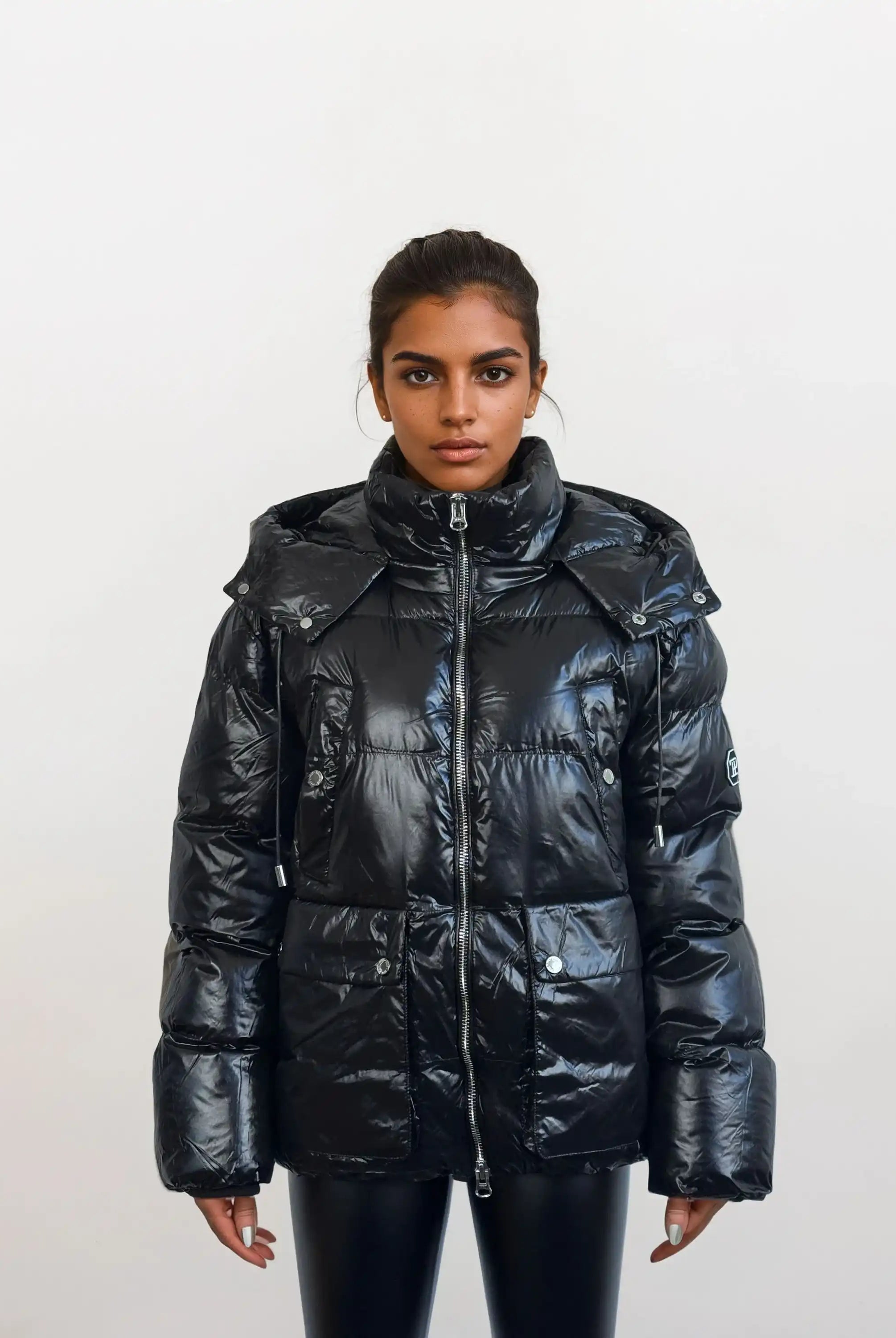 Eine Person steht vor einem weißen Hintergrund und trägt die trueprodigy Damen Winterjacke mit hochglänzender Oberfläche im Regular Fit, mit hohem Kragen, abnehmbarem Kapuze, großen Taschen, sichtbarem Frontreißverschluss und schwarzer Hose.