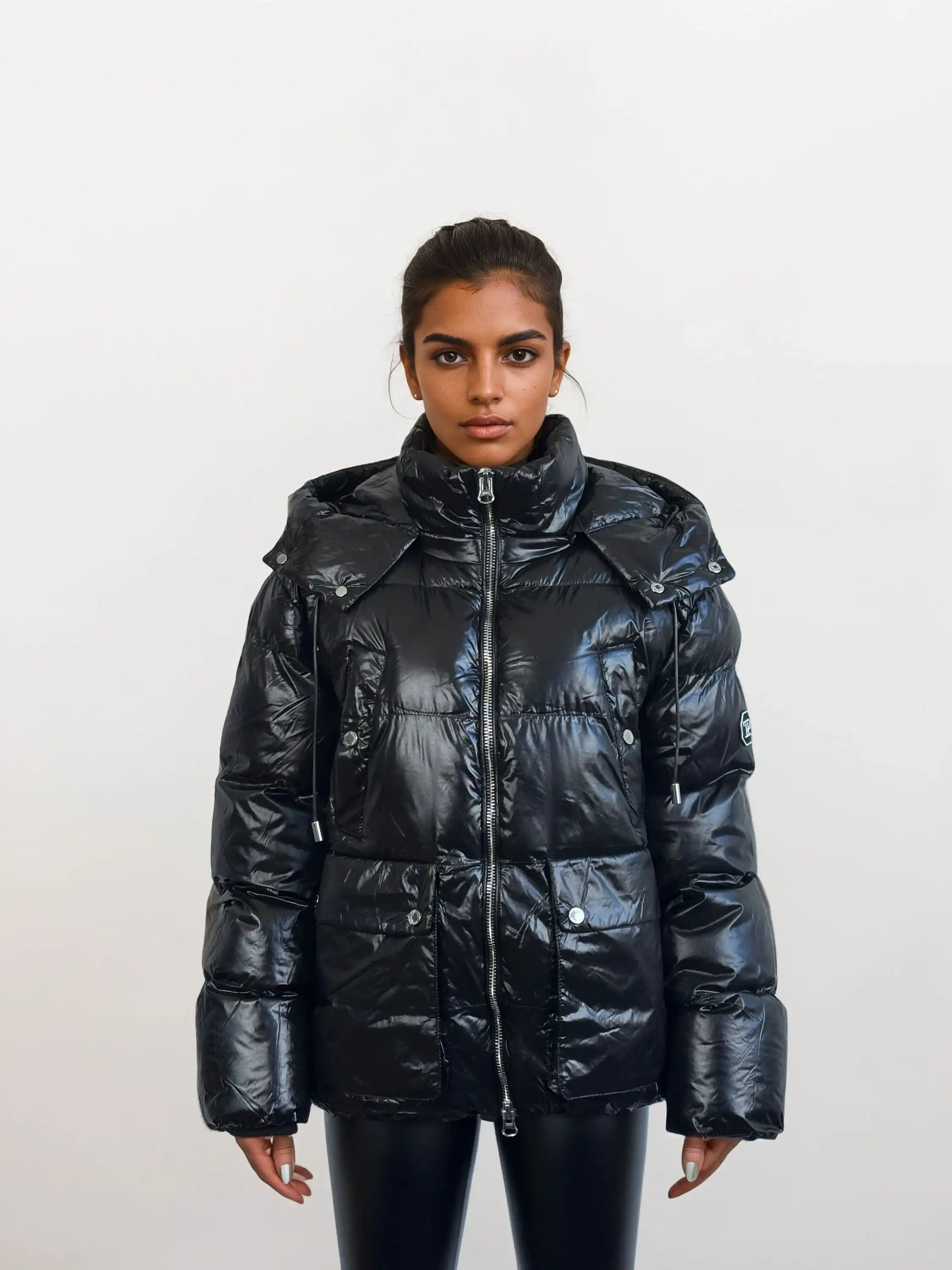 Eine Person steht vor einem weißen Hintergrund und trägt die trueprodigy Damen Winterjacke mit hochglänzender Oberfläche im Regular Fit, mit hohem Kragen, abnehmbarem Kapuze, großen Taschen, sichtbarem Frontreißverschluss und schwarzer Hose.