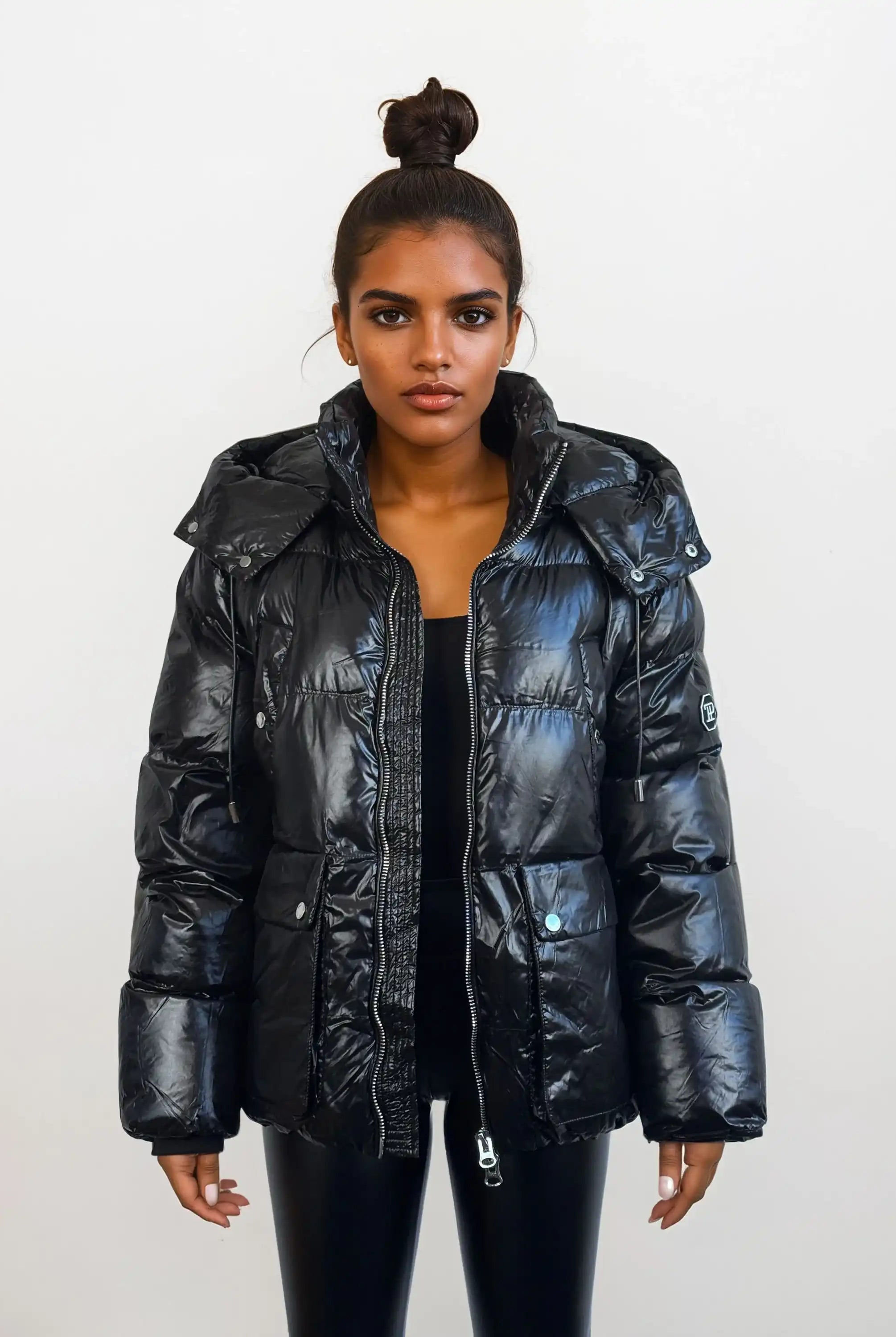 Eine Frau mit dunklem Haar in einem Haarknoten trägt die trueprodigy Damen Winterjacke mit hochglänzender Oberfläche im Regular Fit über einem schwarzen Oberteil und einer Hose vor einem schlichten weißen Hintergrund.