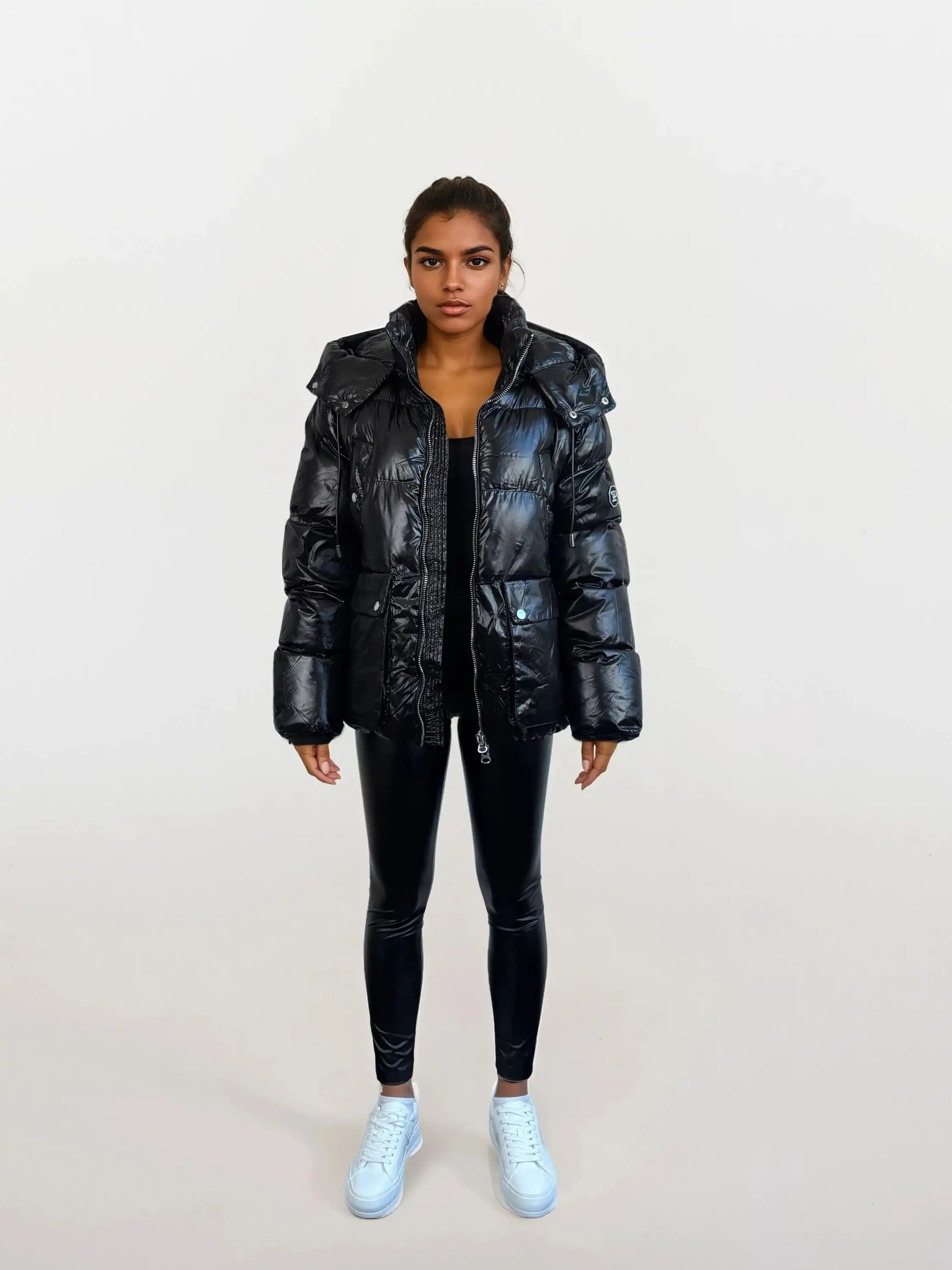 Eine Frau blickt nach vorne in einer trueprodigy Damen Winterjacke mit hochglänzender Oberfläche im Regular Fit, gepaart mit schwarzen Leggings und weißen Sneakers. Sie posiert mit entspannten Händen an den Seiten vor einem hellen, schlichten Hintergrund.