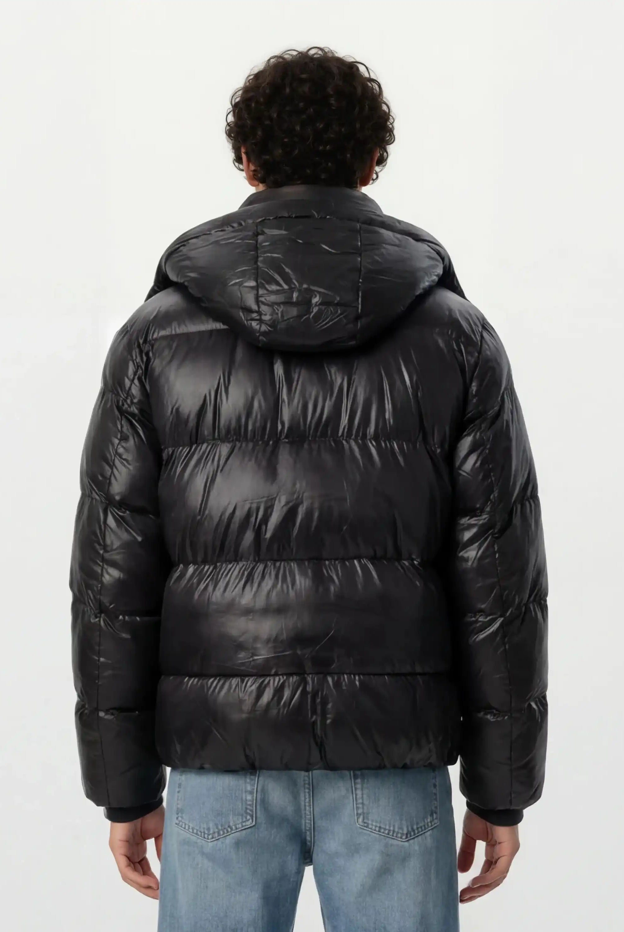 Eine Person mit lockigem dunklem Haar trägt eine trueprodigy Herren Winterjacke mit hochglänzender Oberfläche im Regular Fit und eine hellblaue Jeans, die von der Kamera abgewandt vor einem schlichten weißen Hintergrund steht.