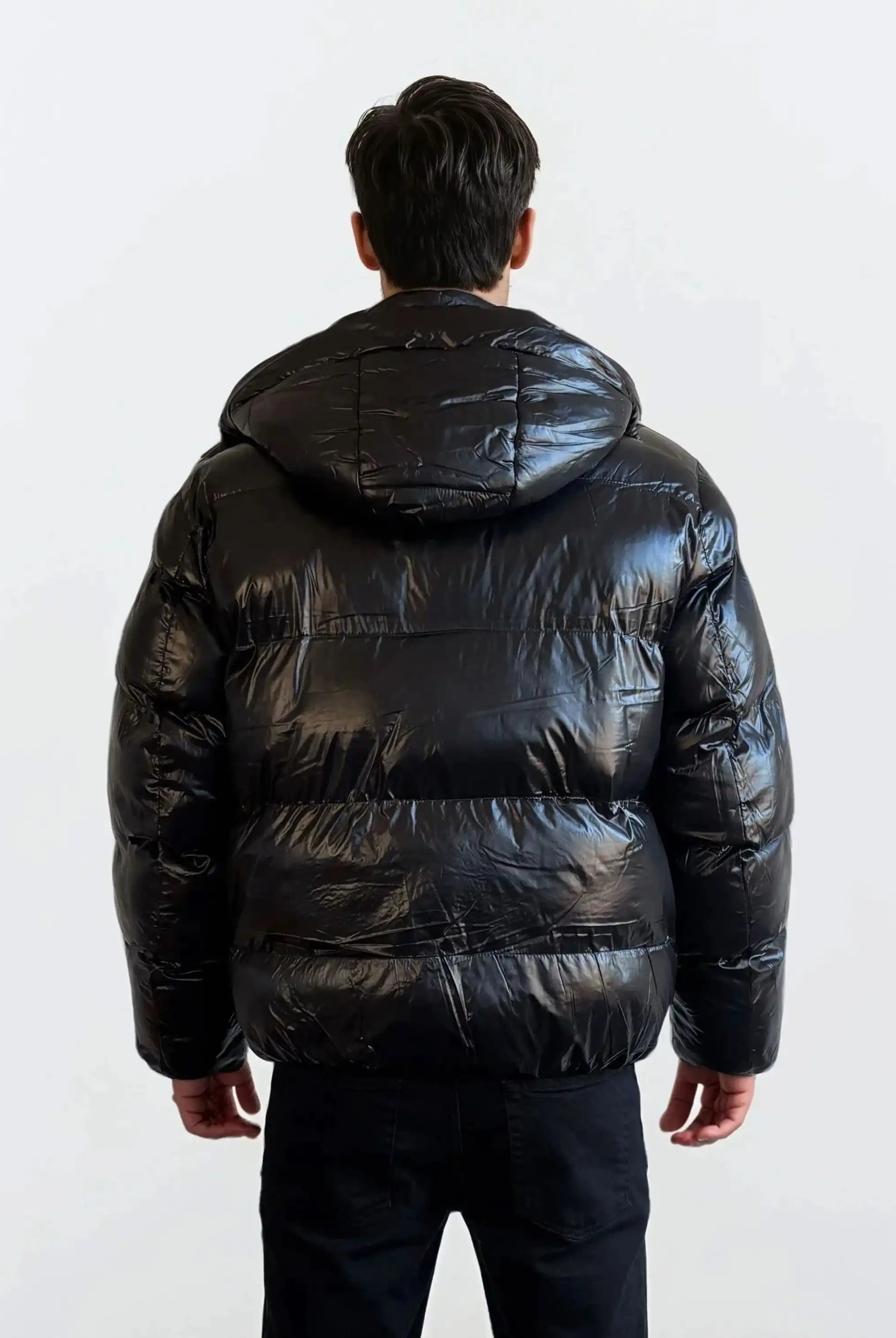Eine Person steht in einer schwarzen Hose und einer trueprodigy Herren Winterjacke mit hochglänzender Oberfläche im Regular Fit mit Kapuze vor einem schlichten hellen Hintergrund vor der Kamera.