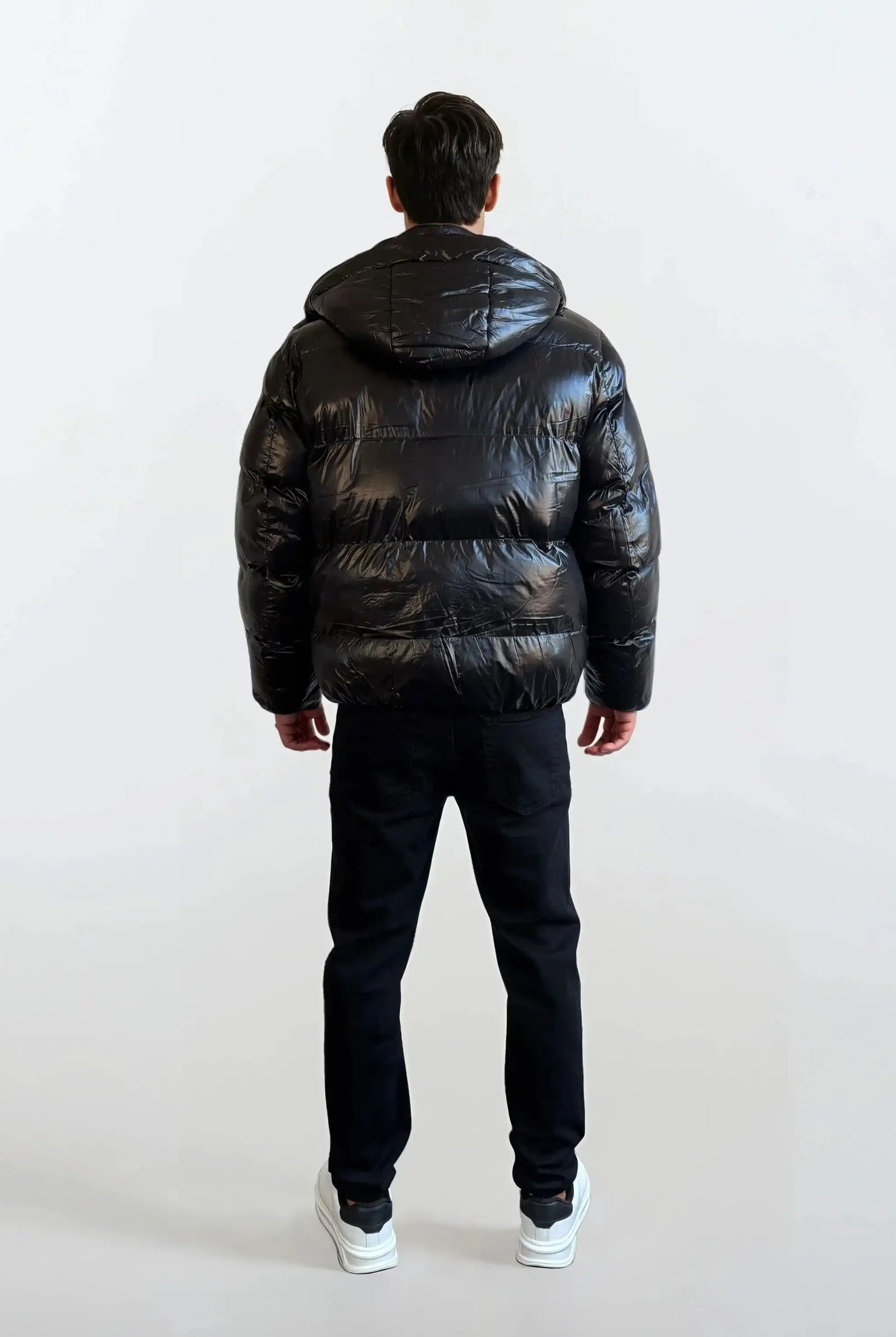 Eine Person in einer trueprodigy Herren Winterjacke mit hochglänzender Oberfläche im Regular Fit, schwarzer Hose und weißen Turnschuhen steht von der Kamera abgewandt vor einem schlichten hellen Hintergrund.