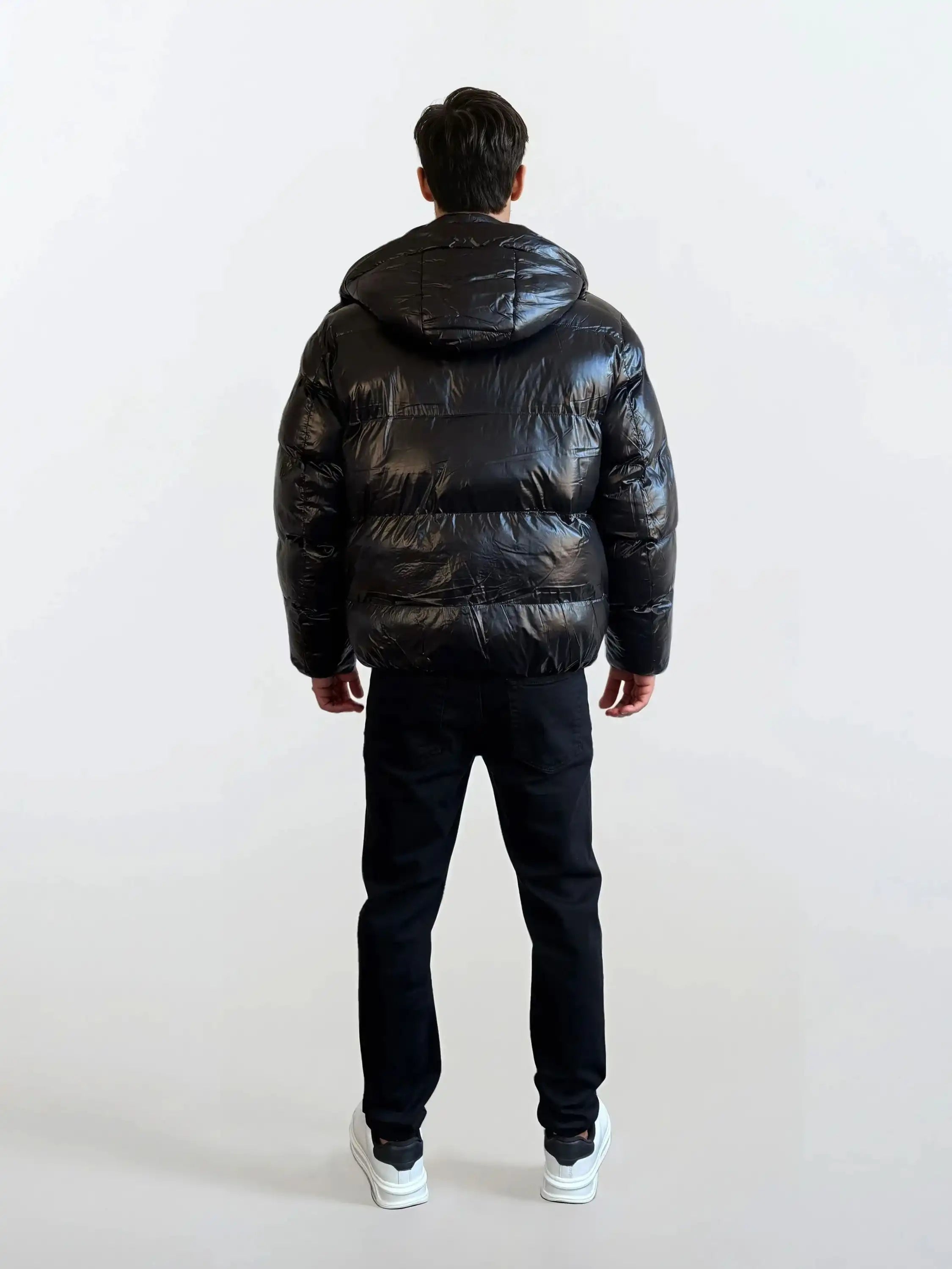 Eine Person in einer trueprodigy Herren Winterjacke mit hochglänzender Oberfläche im Regular Fit, schwarzer Hose und weißen Turnschuhen steht von der Kamera abgewandt vor einem schlichten hellen Hintergrund.