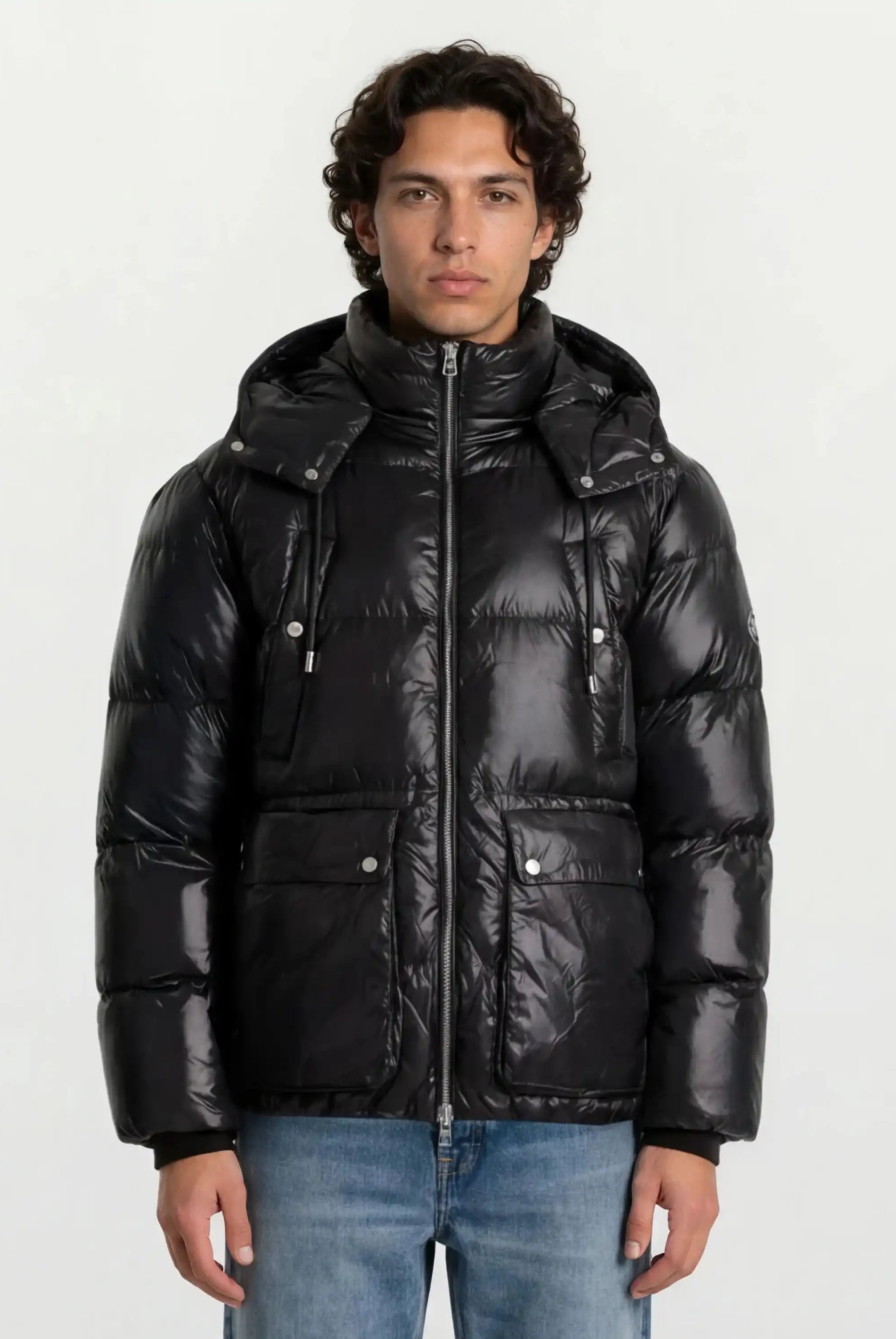 Ein Mann trägt die Herren Winterjacke mit hochglänzender Oberfläche im Regular Fit von trueprodigy mit Kapuze, Frontreißverschluss und zwei großen Knopftaschen sowie hellblauen Jeans vor schlicht weißem Hintergrund.