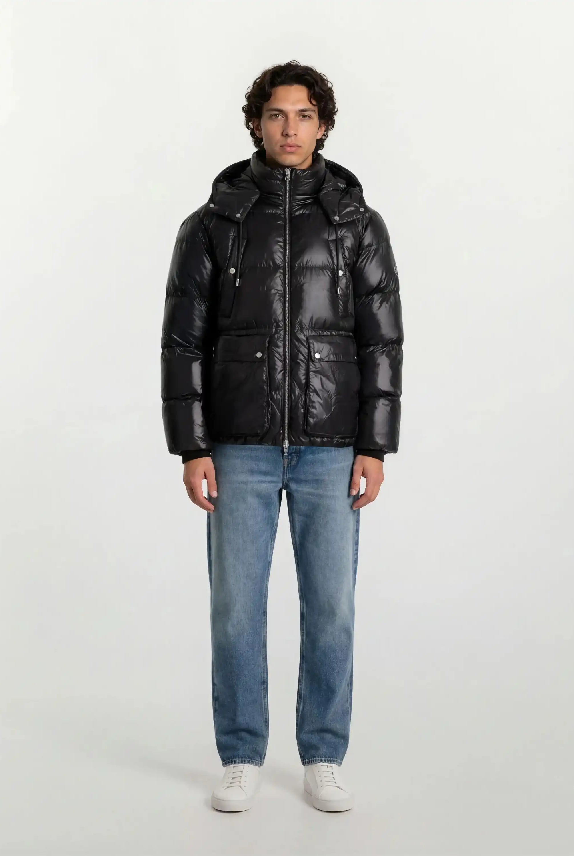 Eine Person steht mit Blick nach vorne in einer trueprodigy Herren Winterjacke mit hochglänzender Oberfläche im Regular Fit, hellblauen Jeans und weißen Turnschuhen vor einem schlichten hellen Hintergrund.