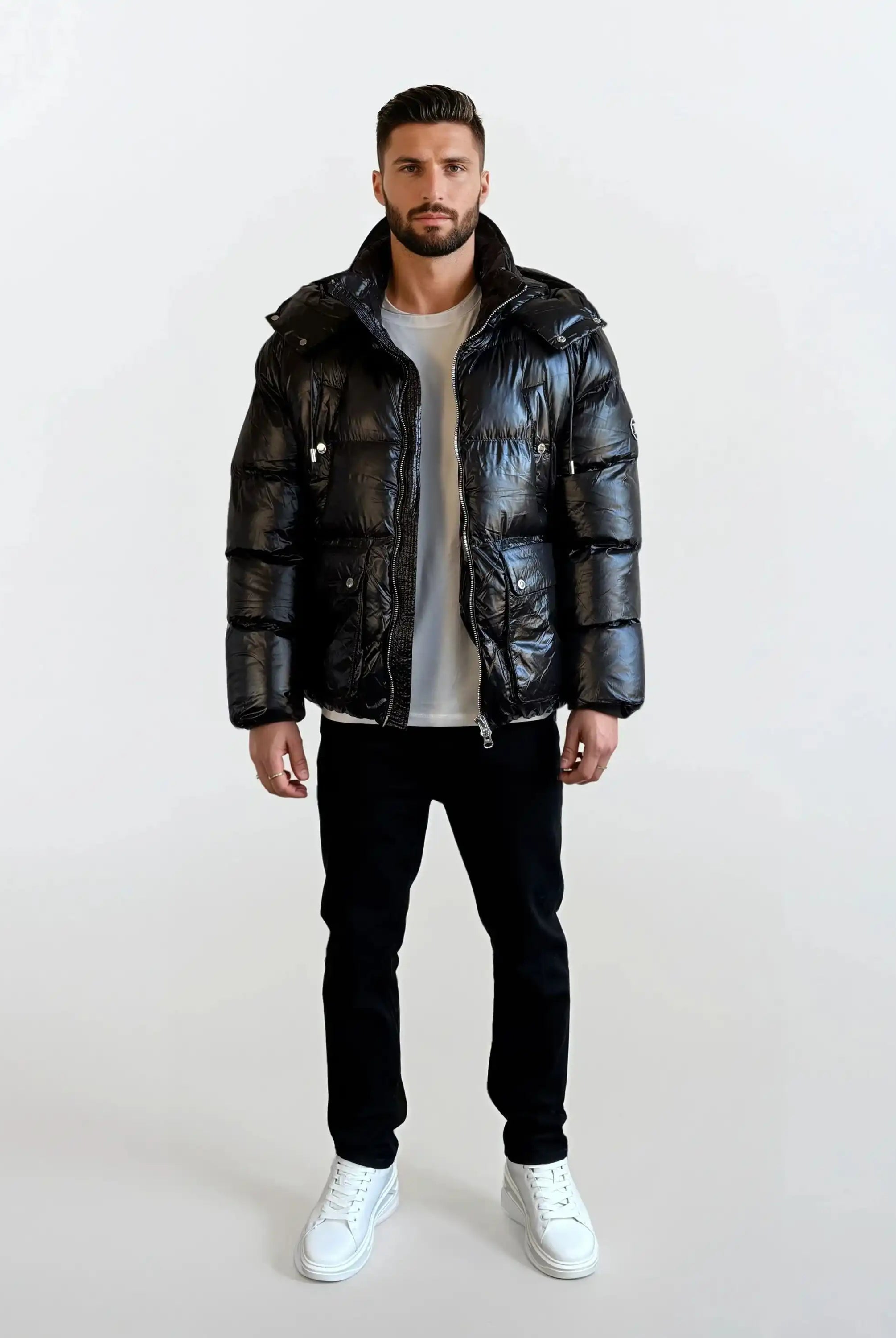 Ein Mann steht mit dem Gesicht nach vorne vor einem schlichten weißen Hintergrund und trägt die trueprodigy Herren Winterjacke mit hochglänzender Oberfläche im Regular Fit, gepaart mit einem weißen T-Shirt, einer schwarzen Hose und weißen Sneakers.