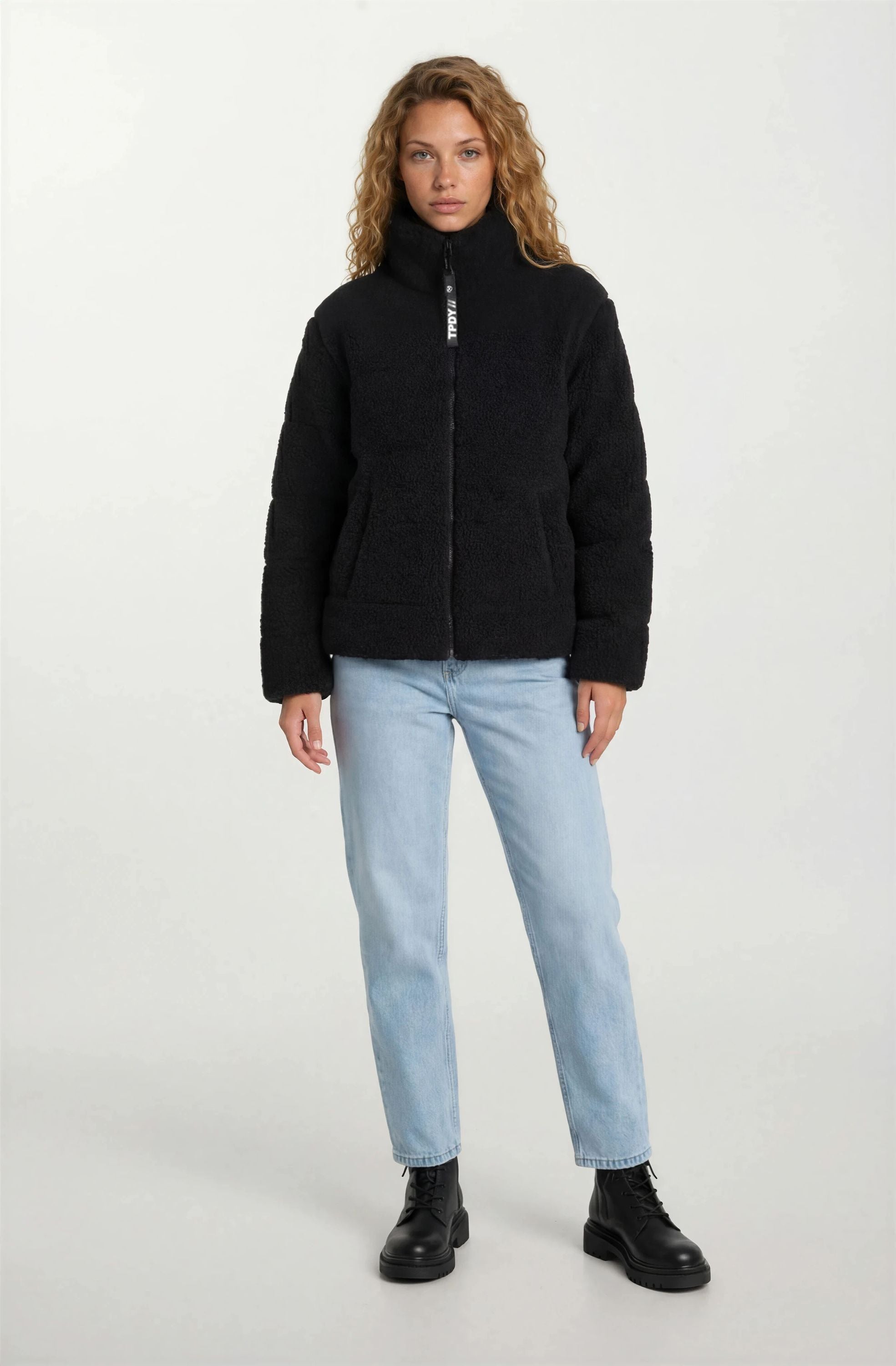 Eine Person steht mit Blick nach vorne und trägt die trueprodigy Damen Winterjacke mit flauschiger Sherpa-Oberfläche im Oversized Fit in Schwarz, gestylt mit hellblauen Jeans und schwarzen Schnürstiefeln vor einem schlichten, hellen Hintergrund.