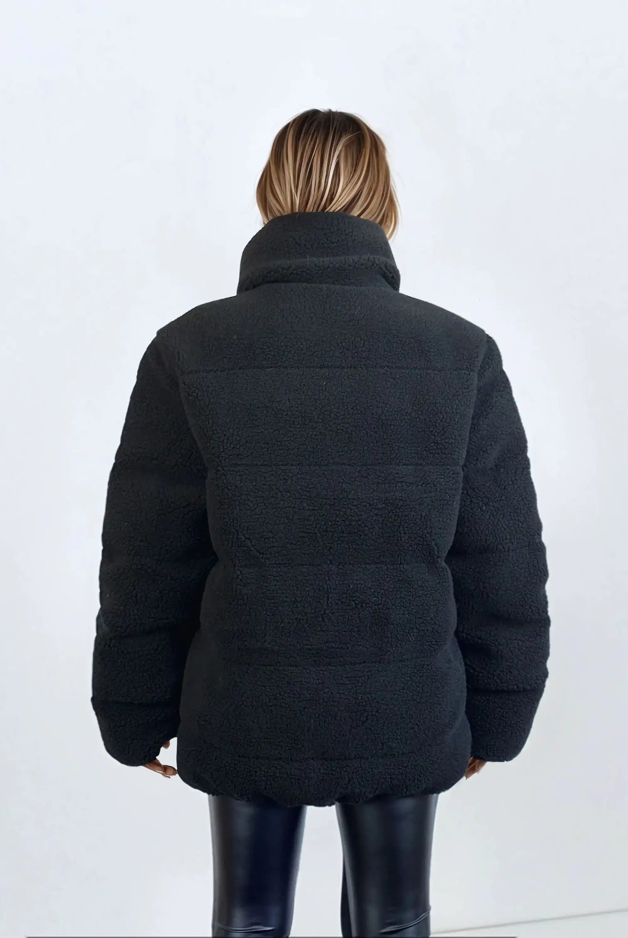 Eine Person mit schulterlangem Haar, von hinten gesehen, trägt eine schwarze Damen Winterjacke mit flauschiger Sherpa-Oberfläche im Oversized Fit von trueprodigy und schwarze Lederhose. Der Hintergrund ist einfarbig und hell.