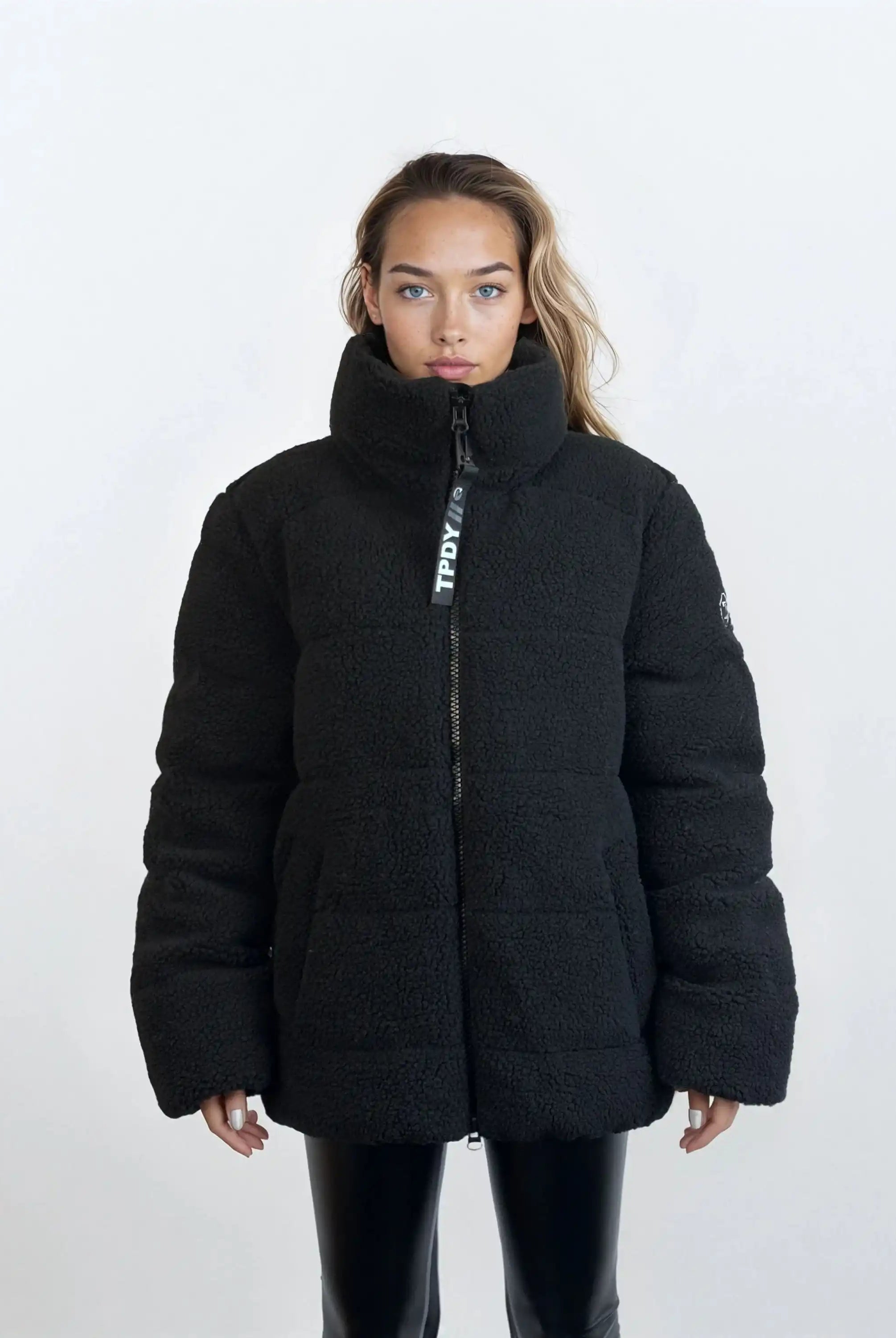 Eine Frau mit langen Haaren trägt eine trueprodigy Damen Winterjacke mit flauschiger Sherpa-Oberfläche im Oversized Fit in Schwarz, kombiniert mit einer schwarzen Hose. Sie steht frontal vor einem schlichten, hellen Hintergrund.