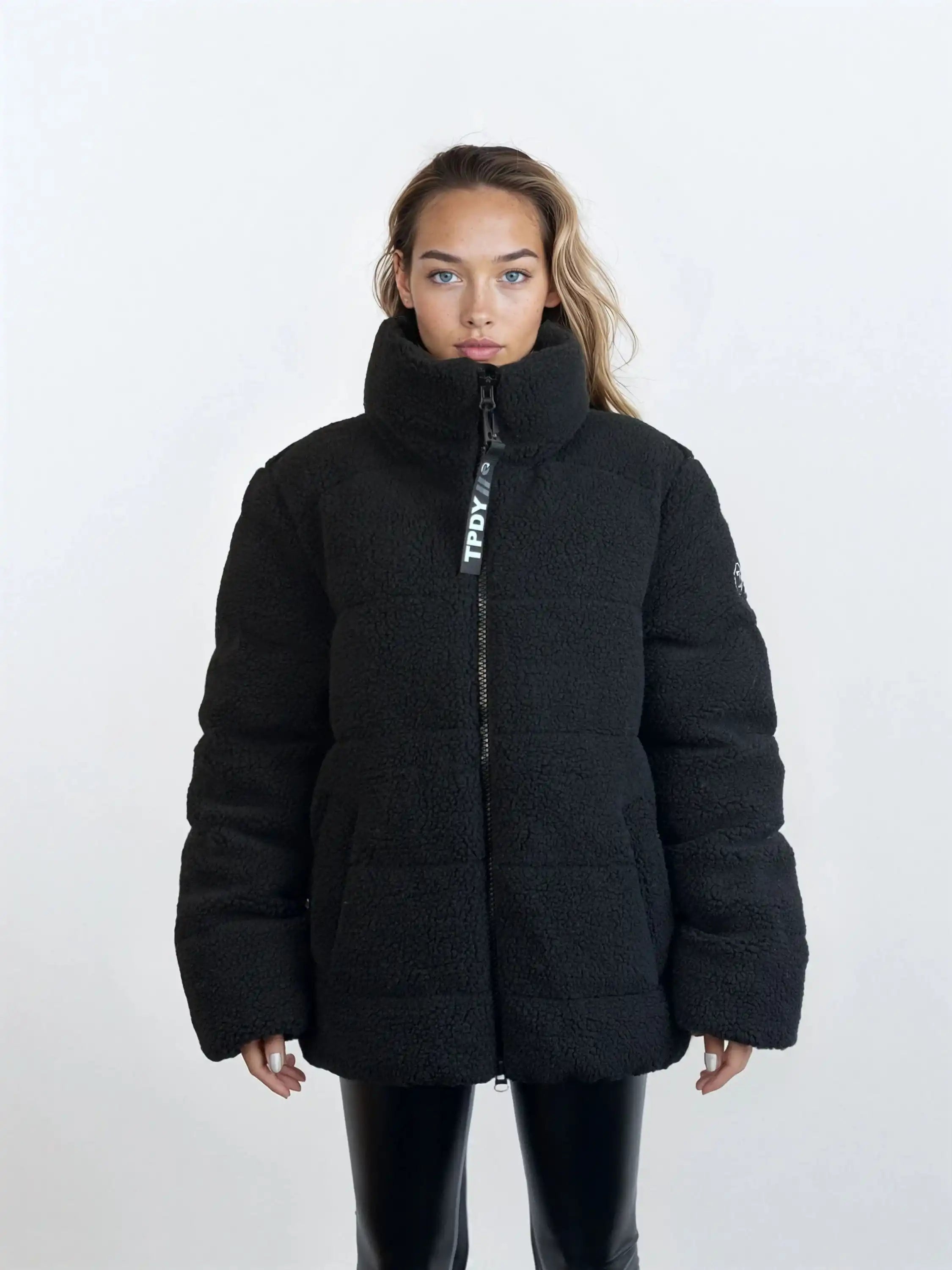 Eine Frau mit langen Haaren trägt eine trueprodigy Damen Winterjacke mit flauschiger Sherpa-Oberfläche im Oversized Fit in Schwarz, kombiniert mit einer schwarzen Hose. Sie steht frontal vor einem schlichten, hellen Hintergrund.
