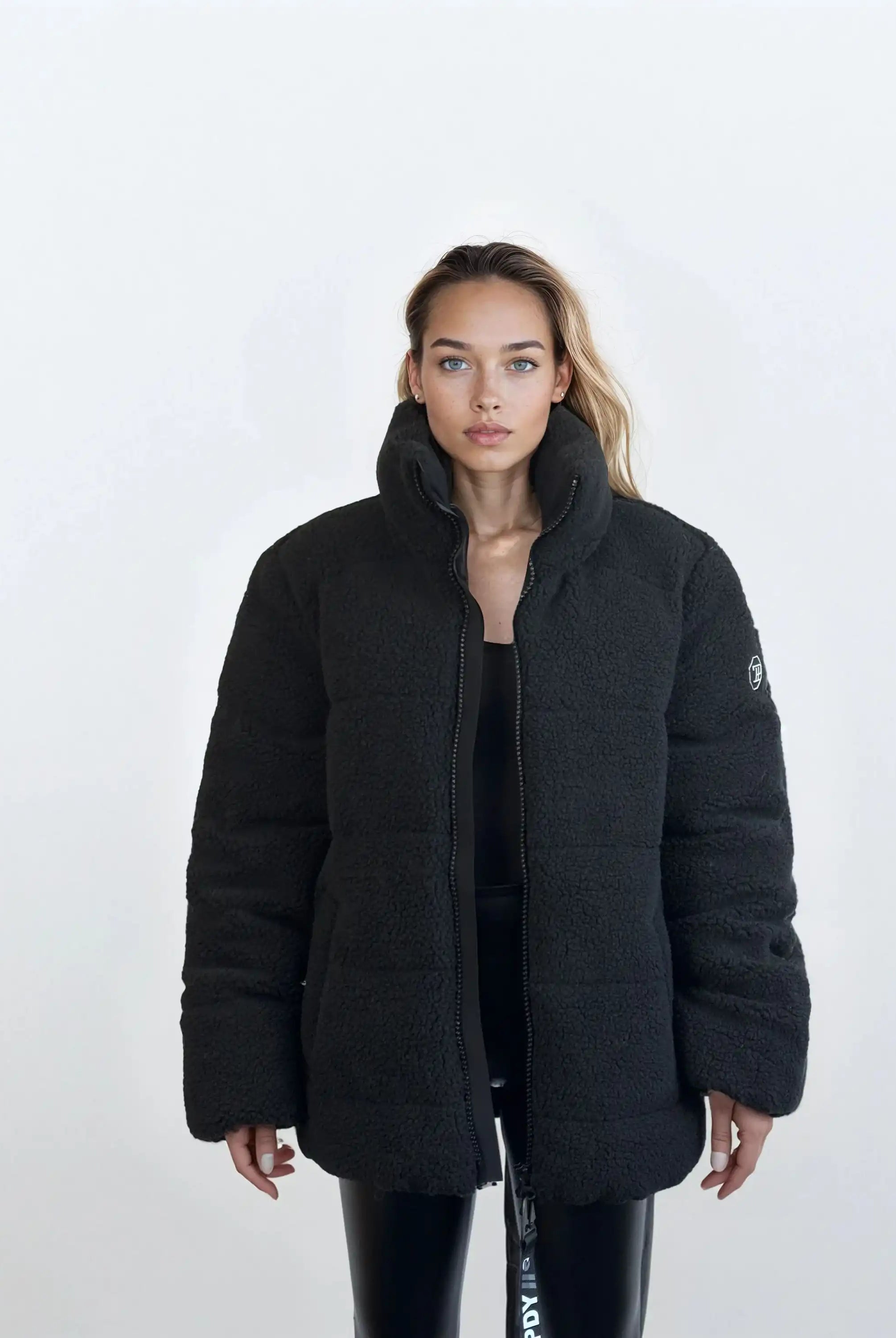 Eine Frau mit langen, hellbraunen Haaren steht vor einem schlichten weißen Hintergrund und trägt die trueprodigy Damen Winterjacke mit flauschiger Sherpa-Oberfläche im Oversized Fit über schwarzer Kleidung. Sie blickt mit neutralem Blick in die Kamera.