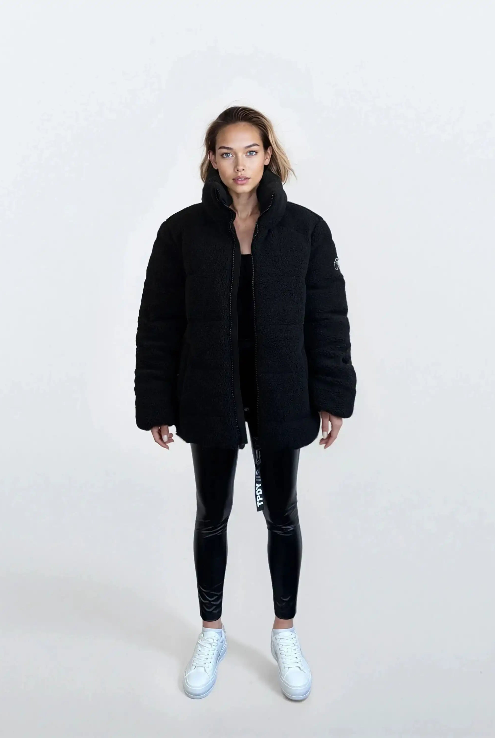 Eine Person steht mit dem Gesicht nach vorne vor einem schlichten, hellen Hintergrund und trägt die trueprodigy Damen Winterjacke mit flauschiger Sherpa-Oberfläche im Oversized Fit, schwarz glänzende Leggings und weiße Turnschuhe.