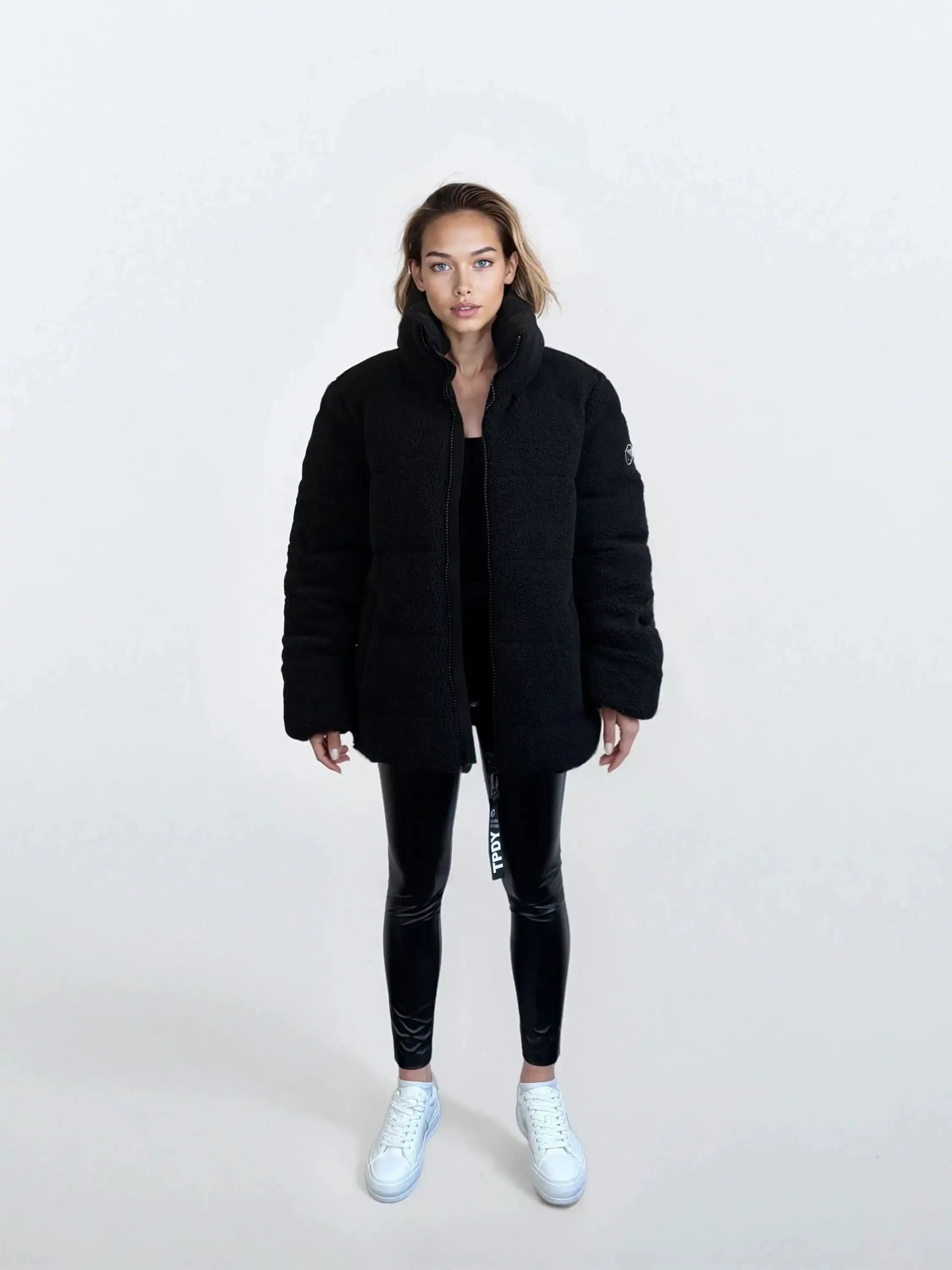 Eine Person steht mit dem Gesicht nach vorne vor einem schlichten, hellen Hintergrund und trägt die trueprodigy Damen Winterjacke mit flauschiger Sherpa-Oberfläche im Oversized Fit, schwarz glänzende Leggings und weiße Turnschuhe.
