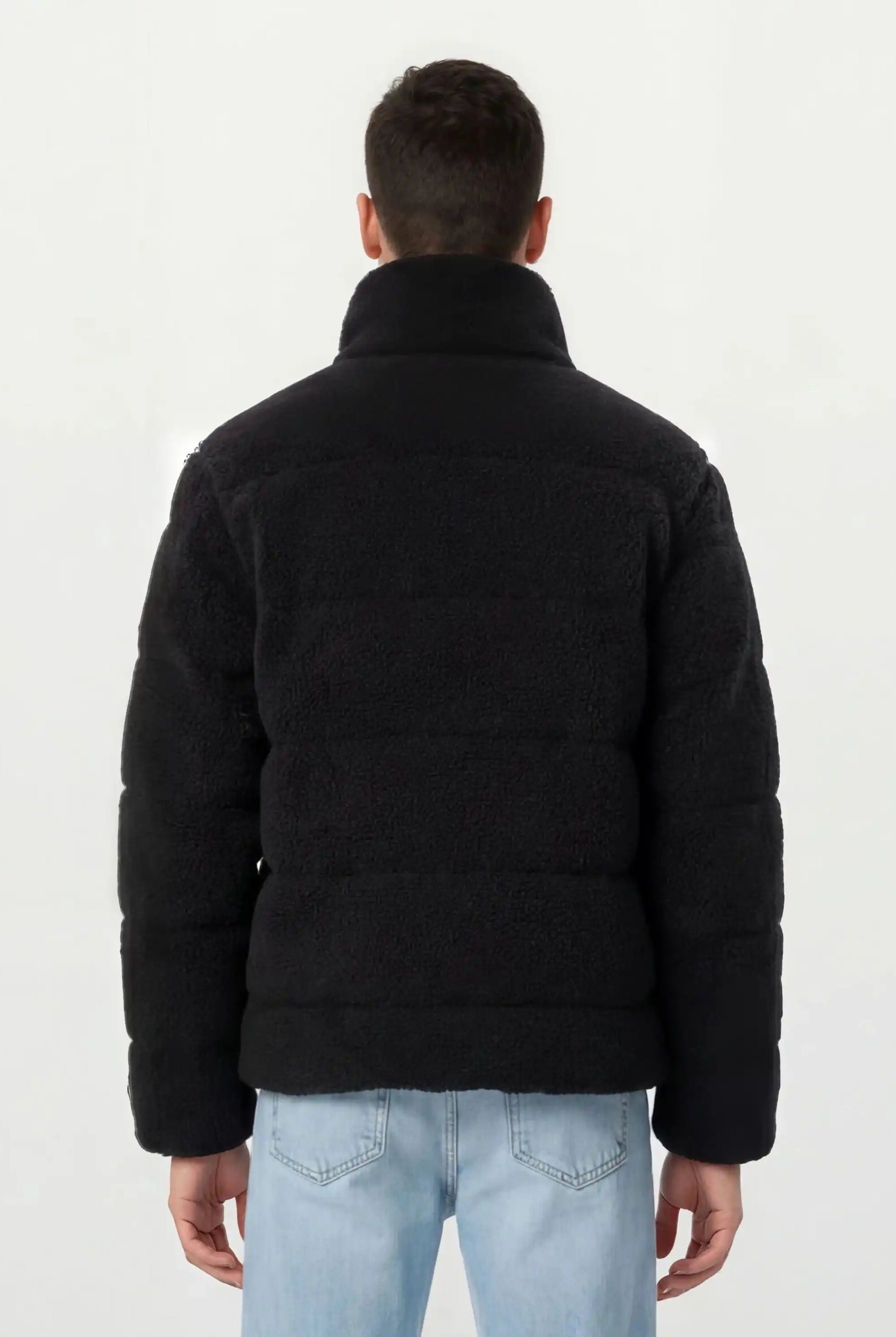 Eine Person wird von hinten gezeigt, die eine schwarze, trueprodigy Herren Winterjacke mit flauschiger Sherpa-Oberfläche im Oversized Fit und hellblaue Jeans vor einem schlichten hellen Hintergrund trägt.