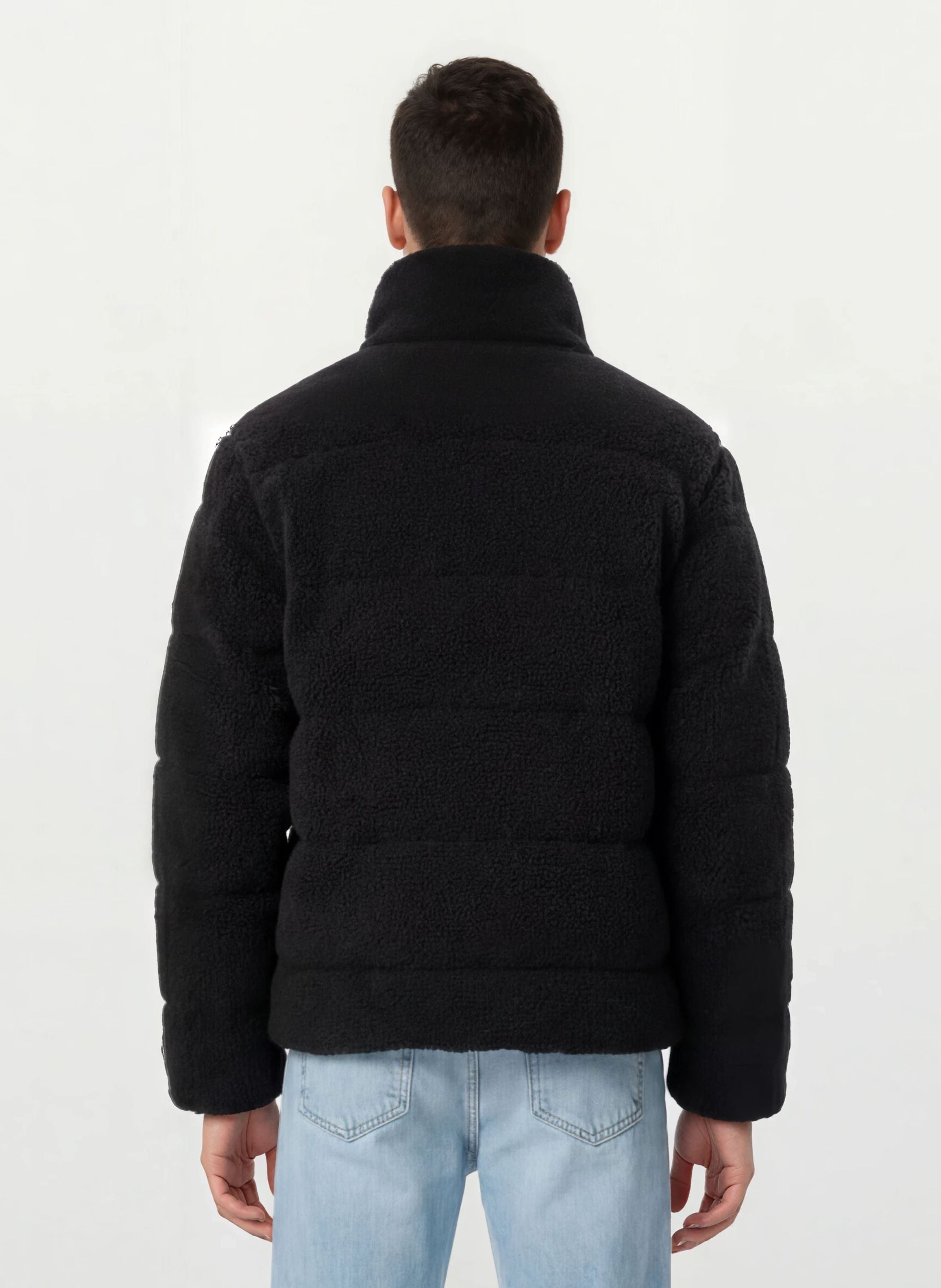 Eine Person wird von hinten gezeigt, die eine schwarze, trueprodigy Herren Winterjacke mit flauschiger Sherpa-Oberfläche im Oversized Fit und hellblaue Jeans vor einem schlichten hellen Hintergrund trägt.