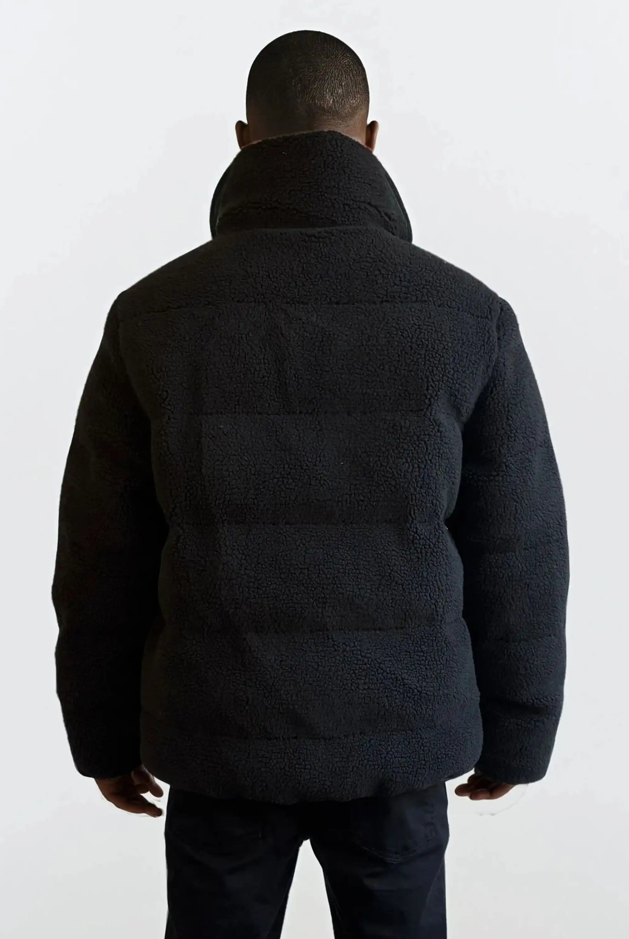 Eine Person steht mit dem Rücken zur Kamera und trägt eine schwarze trueprodigy Herren Winterjacke mit flauschiger Sherpa-Oberfläche im Oversized Fit und eine dunkle Hose vor einem schlichten hellen Hintergrund.
