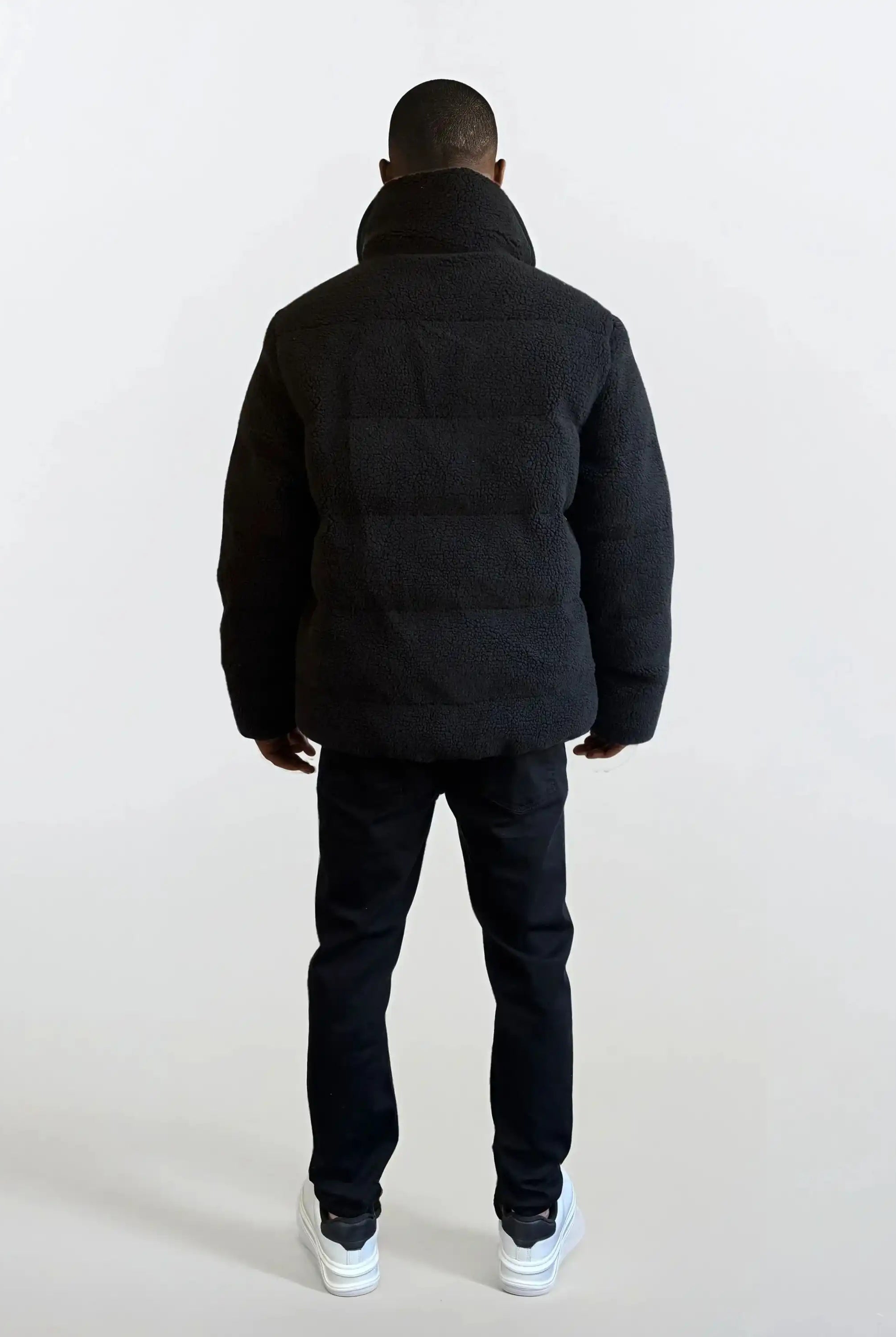 Eine Person schaut von der Kamera weg und trägt eine trueprodigy Herren Winterjacke mit flauschiger Sherpa-Oberfläche im Oversized Fit, eine schwarze Hose und weiße Turnschuhe vor einem schlichten hellen Hintergrund.