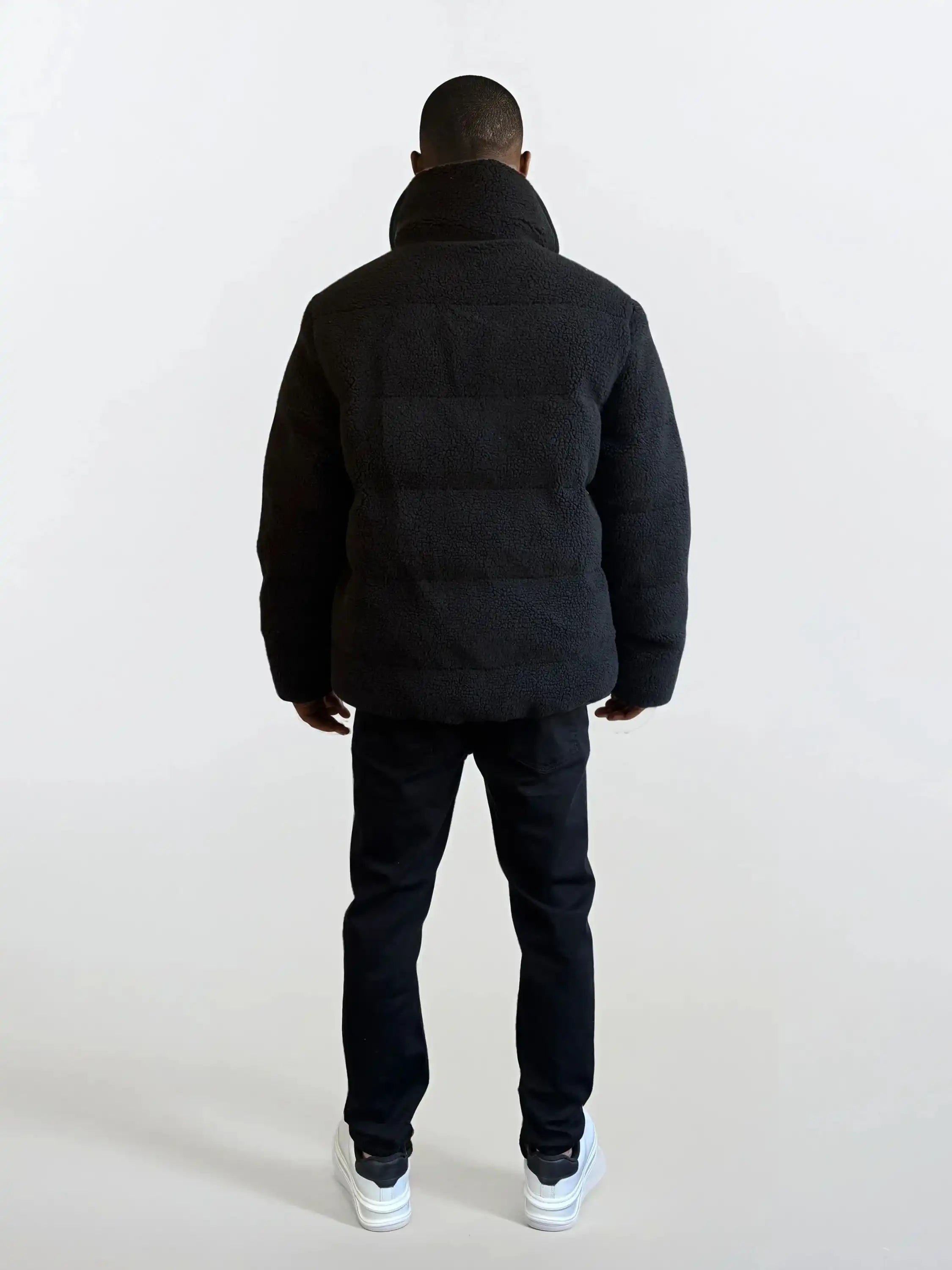 Eine Person schaut von der Kamera weg und trägt eine trueprodigy Herren Winterjacke mit flauschiger Sherpa-Oberfläche im Oversized Fit, eine schwarze Hose und weiße Turnschuhe vor einem schlichten hellen Hintergrund.