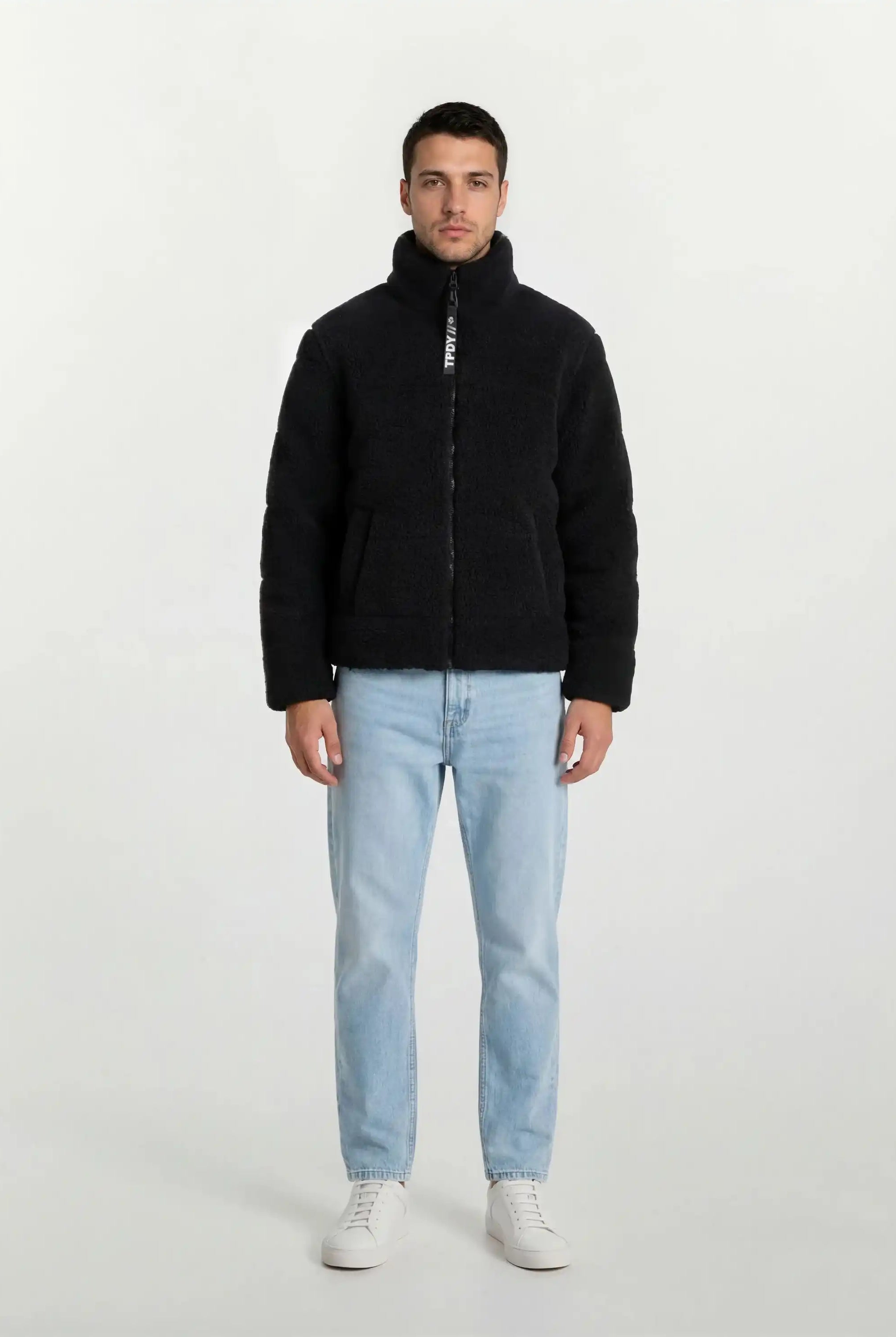 Ein Mann steht mit dem Gesicht nach vorne vor einem schlichten weißen Hintergrund und trägt eine schwarze trueprodigy Herren Winterjacke mit flauschiger Sherpa-Oberfläche im Oversized Fit, hellblaue Jeans und weiße Turnschuhe.