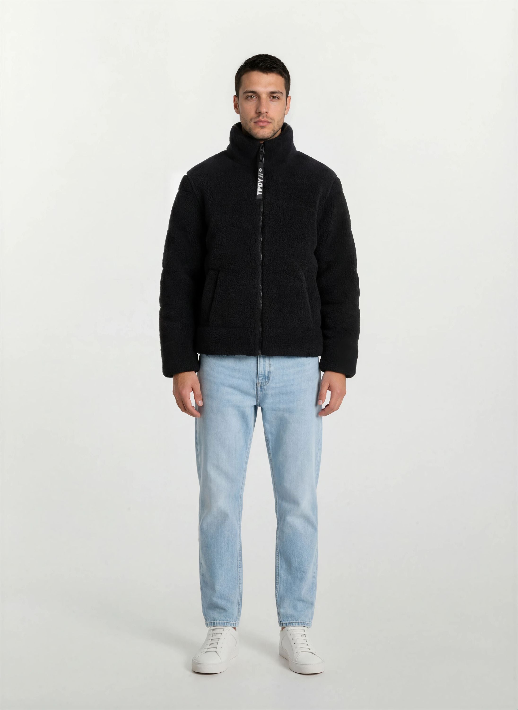 Ein Mann steht mit dem Gesicht nach vorne vor einem schlichten weißen Hintergrund und trägt eine schwarze trueprodigy Herren Winterjacke mit flauschiger Sherpa-Oberfläche im Oversized Fit, hellblaue Jeans und weiße Turnschuhe.