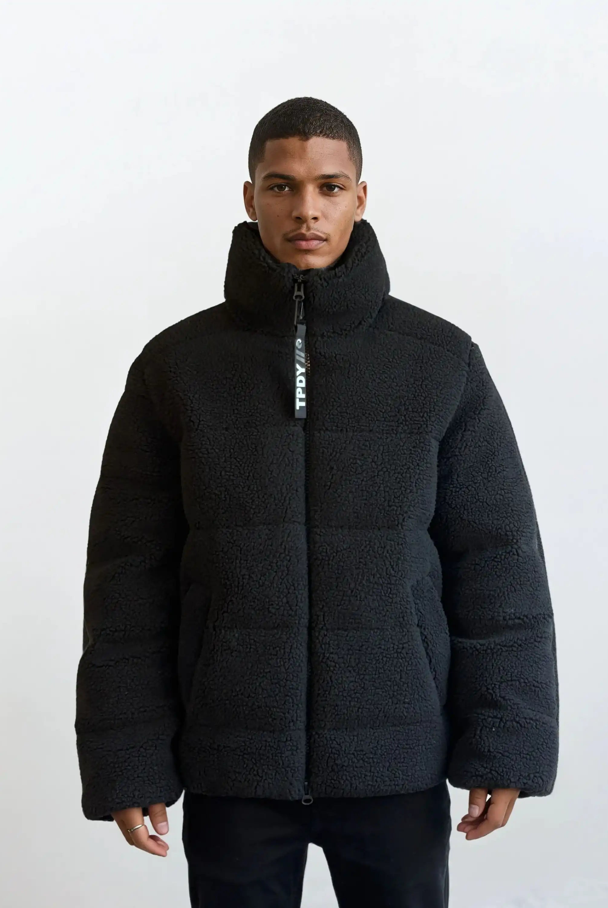 Eine Person steht mit dem Gesicht nach vorne vor einem schlichten weißen Hintergrund und trägt die trueprodigy Herren Winterjacke mit flauschiger Sherpa-Oberfläche im Oversized Fit mit sichtbarem vertikalen Reißverschluss und schwarzer Hose.