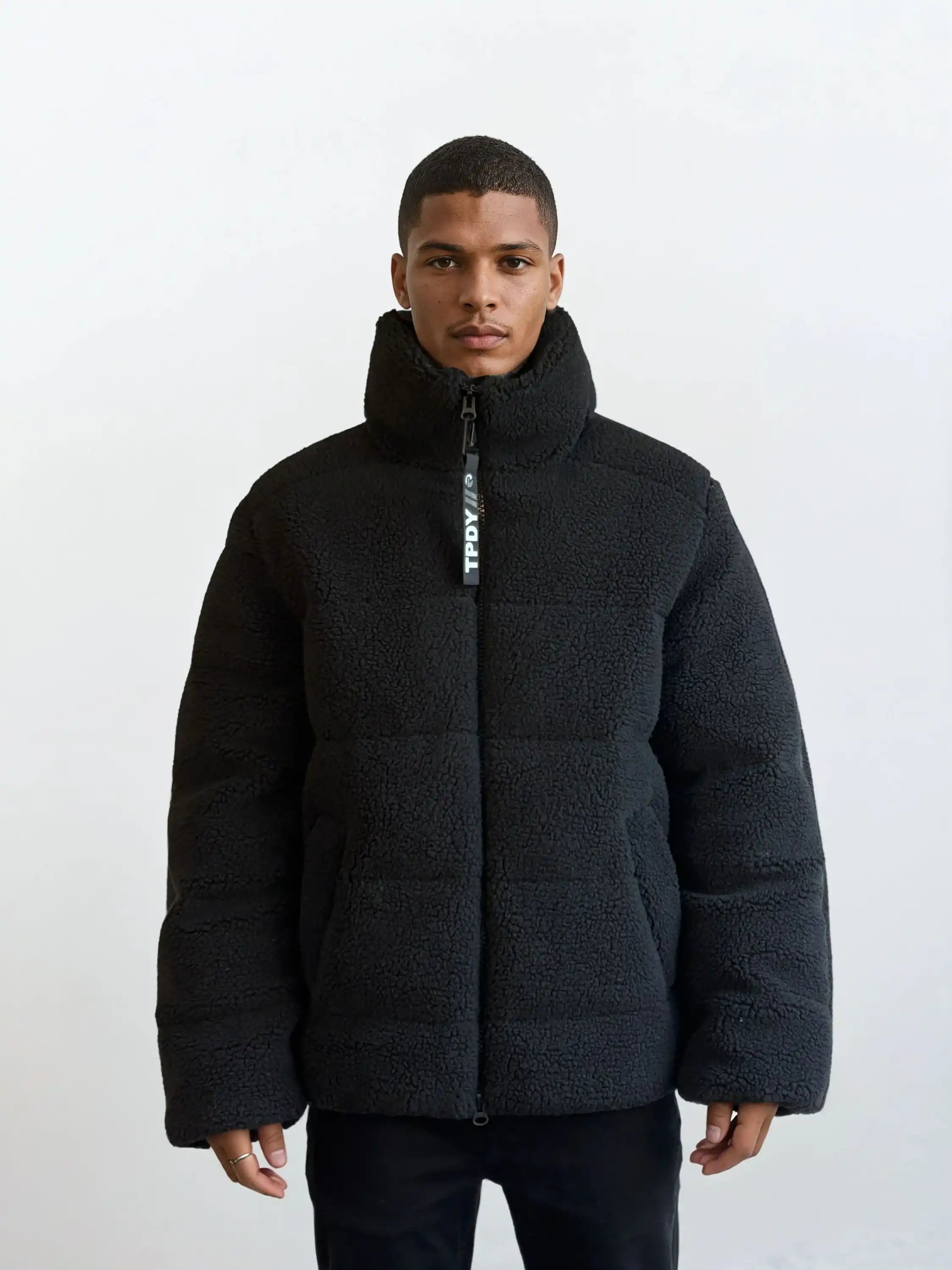Eine Person steht mit dem Gesicht nach vorne vor einem schlichten weißen Hintergrund und trägt die trueprodigy Herren Winterjacke mit flauschiger Sherpa-Oberfläche im Oversized Fit mit sichtbarem vertikalen Reißverschluss und schwarzer Hose.
