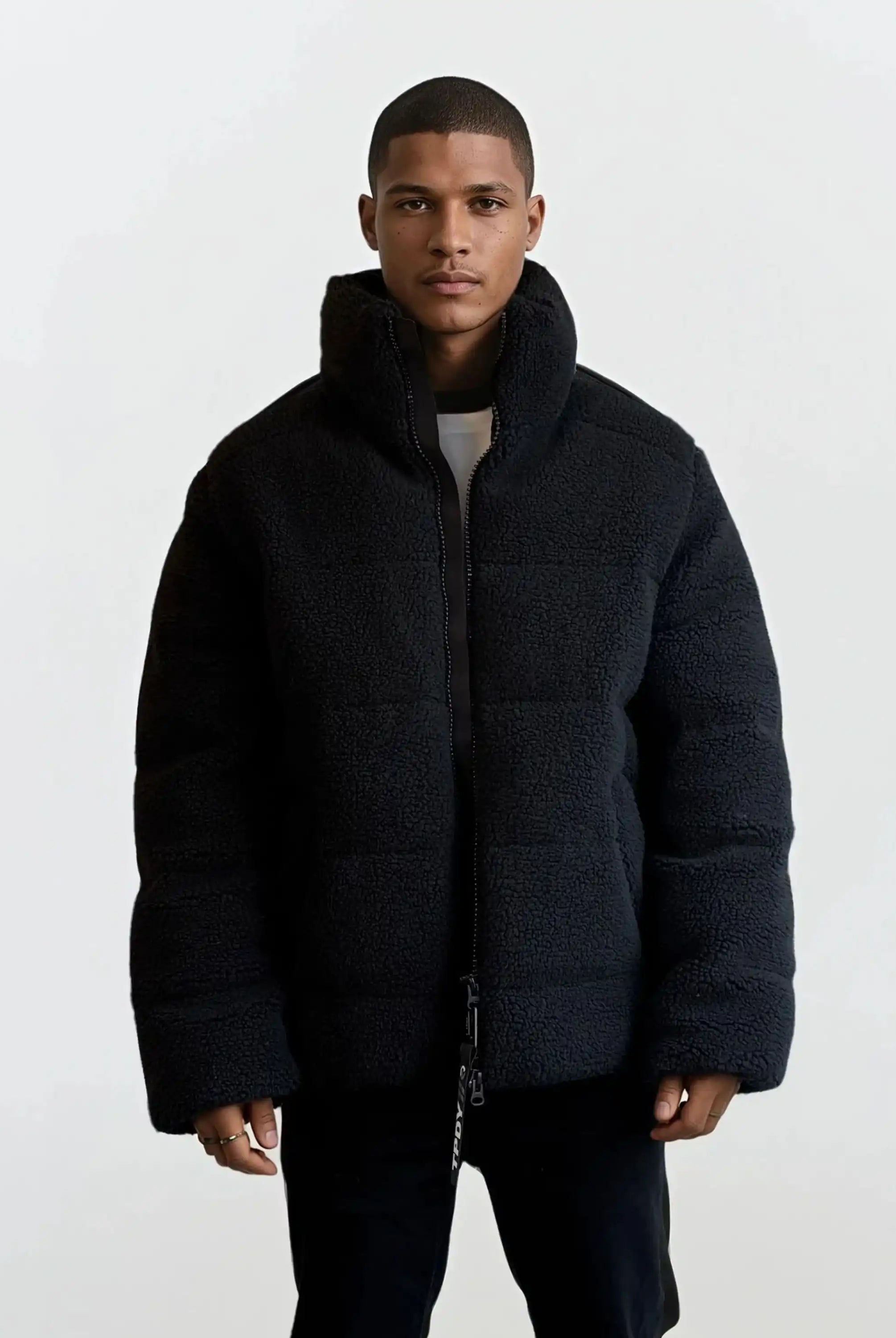 Eine Person steht mit dem Gesicht nach vorne in einer trueprodigy Herren Winterjacke mit flauschiger Sherpa-Oberfläche im Oversized Fit-schwarz und bauschig-über einem weißen Hemd und einer dunklen Hose, vor einem schlichten hellen Hintergrund.