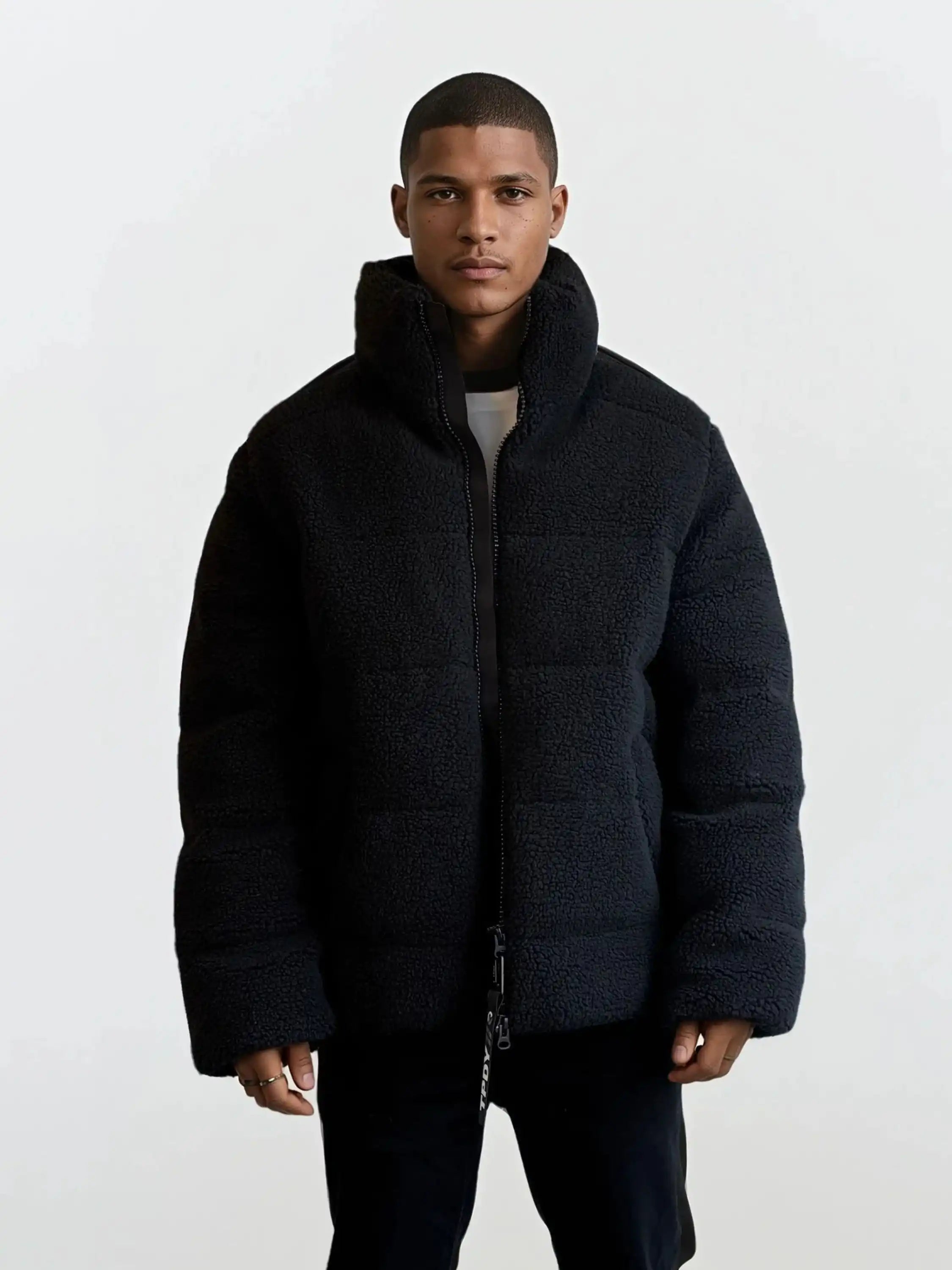Eine Person steht mit dem Gesicht nach vorne in einer trueprodigy Herren Winterjacke mit flauschiger Sherpa-Oberfläche im Oversized Fit-schwarz und bauschig-über einem weißen Hemd und einer dunklen Hose, vor einem schlichten hellen Hintergrund.
