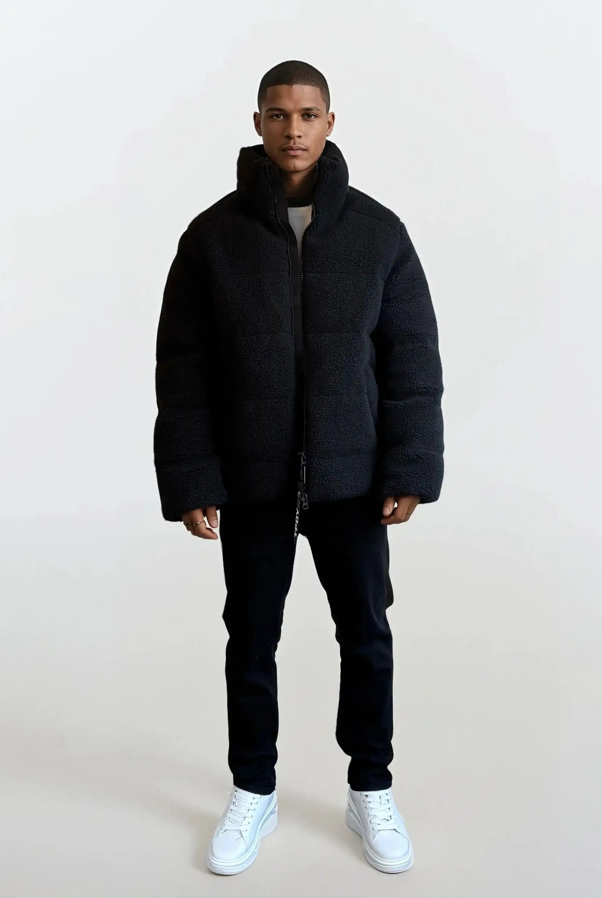 Ein Mann blickt nach vorne und trägt eine schwarze trueprodigy Herren Winterjacke mit flauschiger Sherpa-Oberfläche im Oversized Fit, eine dunkle Hose und weiße Turnschuhe. Der Hintergrund ist einfarbig und hell gefärbt.