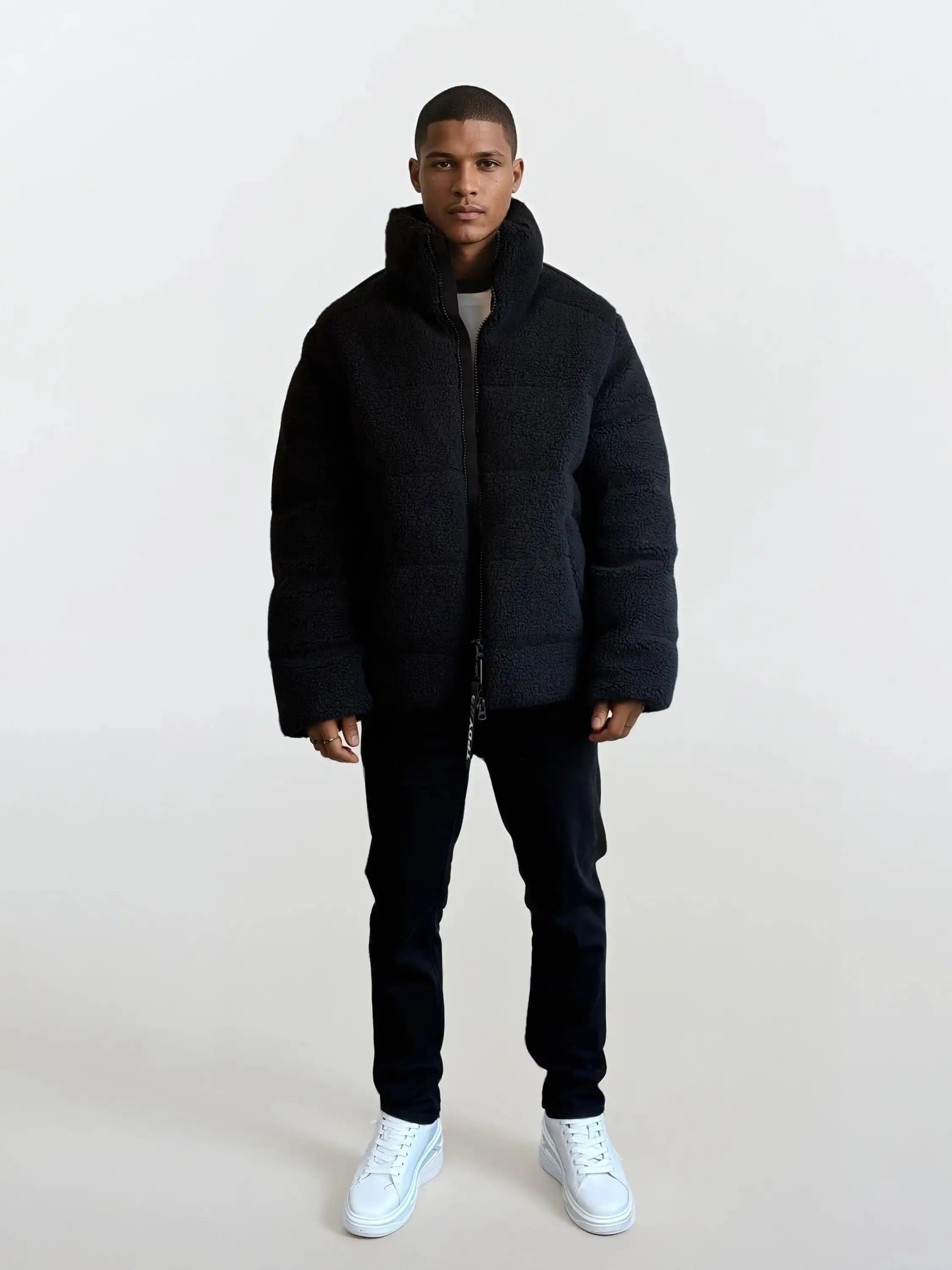Ein Mann blickt nach vorne und trägt eine schwarze trueprodigy Herren Winterjacke mit flauschiger Sherpa-Oberfläche im Oversized Fit, eine dunkle Hose und weiße Turnschuhe. Der Hintergrund ist einfarbig und hell gefärbt.