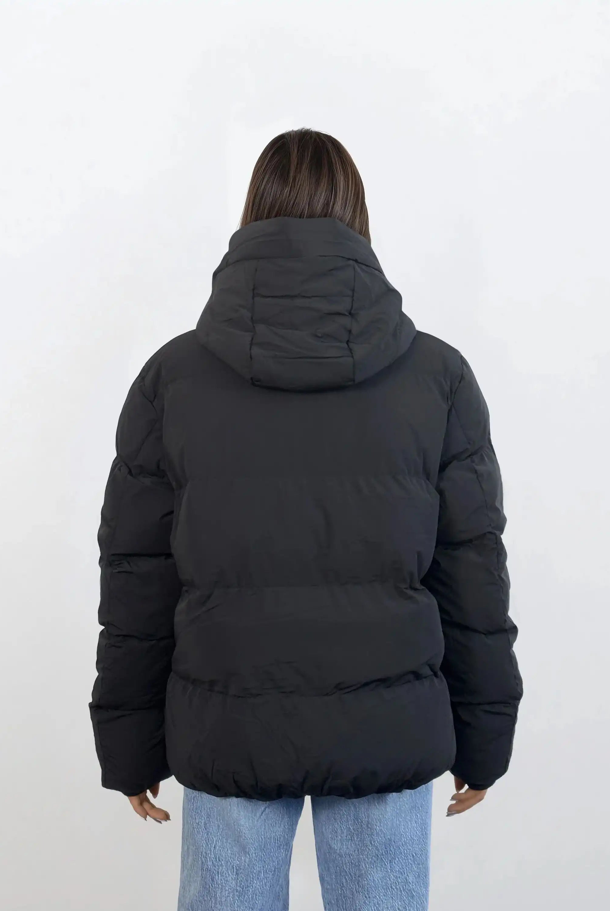 Eine Person mit glattem braunem Haar, von hinten vor einem weißen Hintergrund dargestellt, trägt eine schwarze trueprodigy Damen Winterjacke mit fester Kapuze im Oversized Fit und hellblaue Jeans.