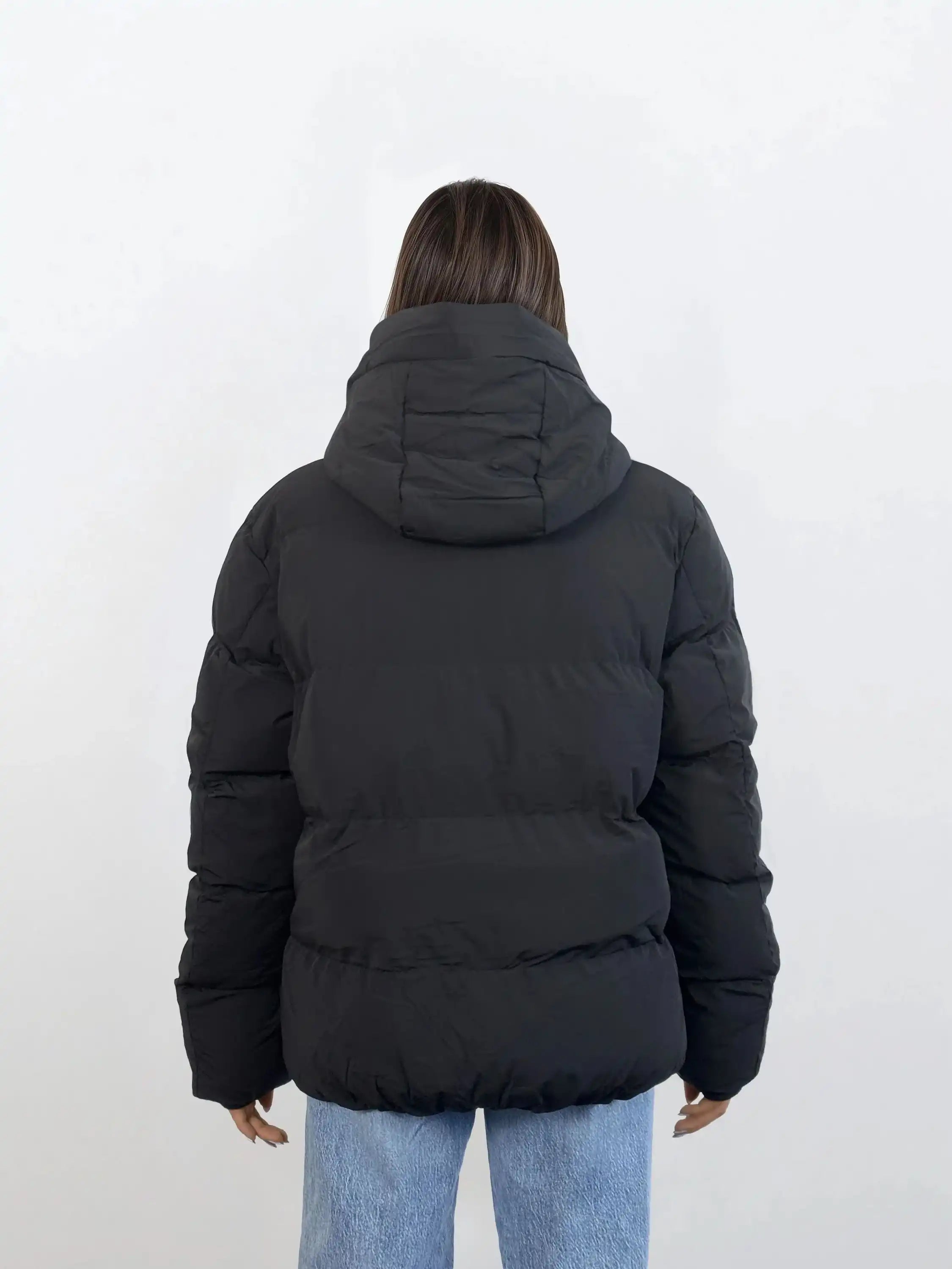 Eine Person mit glattem braunem Haar, von hinten vor einem weißen Hintergrund dargestellt, trägt eine schwarze trueprodigy Damen Winterjacke mit fester Kapuze im Oversized Fit und hellblaue Jeans.