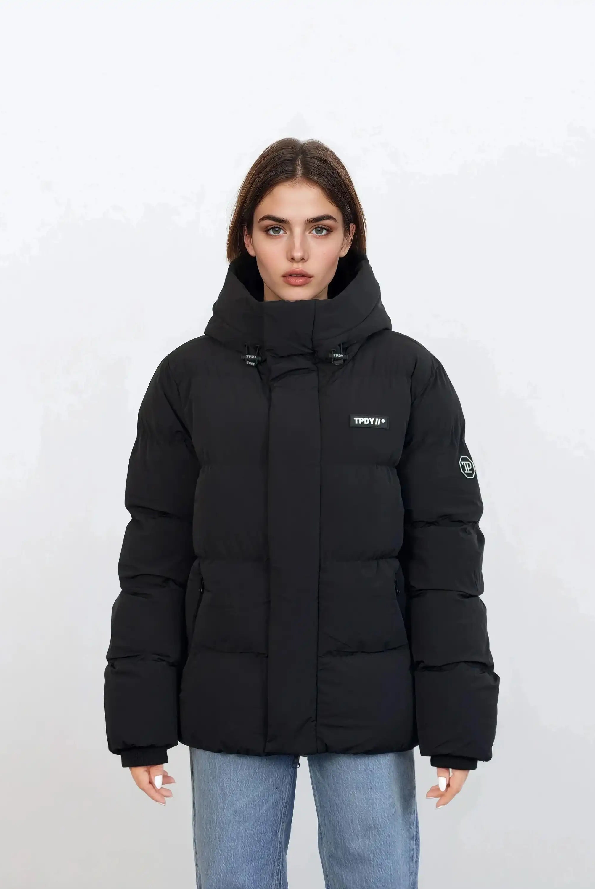 Eine Person mit glattem braunem Haar trägt eine trueprodigy Damen Winterjacke mit fester Kapuze im Oversized Fit in Schwarz mit Logoaufnähern auf der Brust und dem oberen Ärmel, gepaart mit hellblauen Jeans vor einem schlichten weißen Hintergrund.
