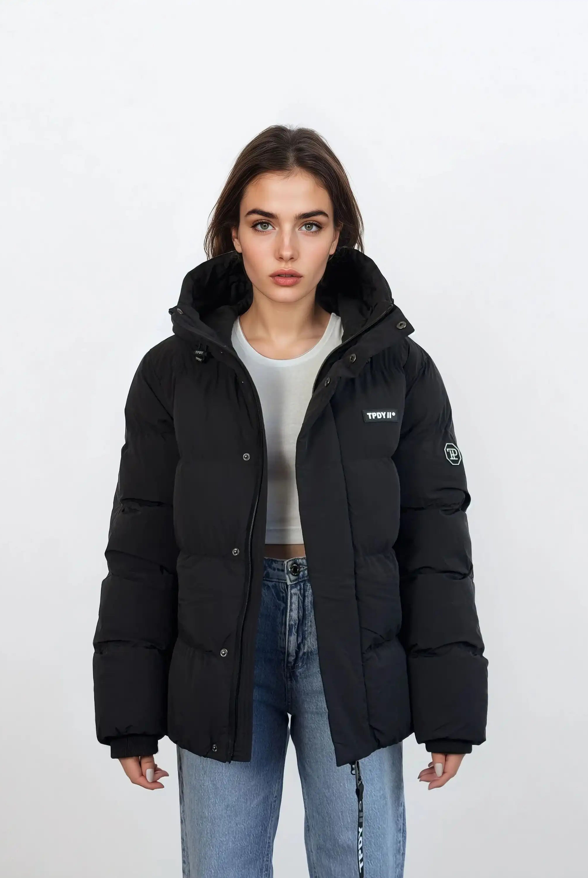 Eine Person steht vor weißem Hintergrund und trägt eine schwarze, wasserabweisende trueprodigy Damen Winterjacke mit fester Kapuze im Oversized Fit über einem hellgrauen Shirt und blauen Jeans. Die Jacke hat einen Aufnäher am Ärmel und den Schriftzug STOFFE auf der Brust.