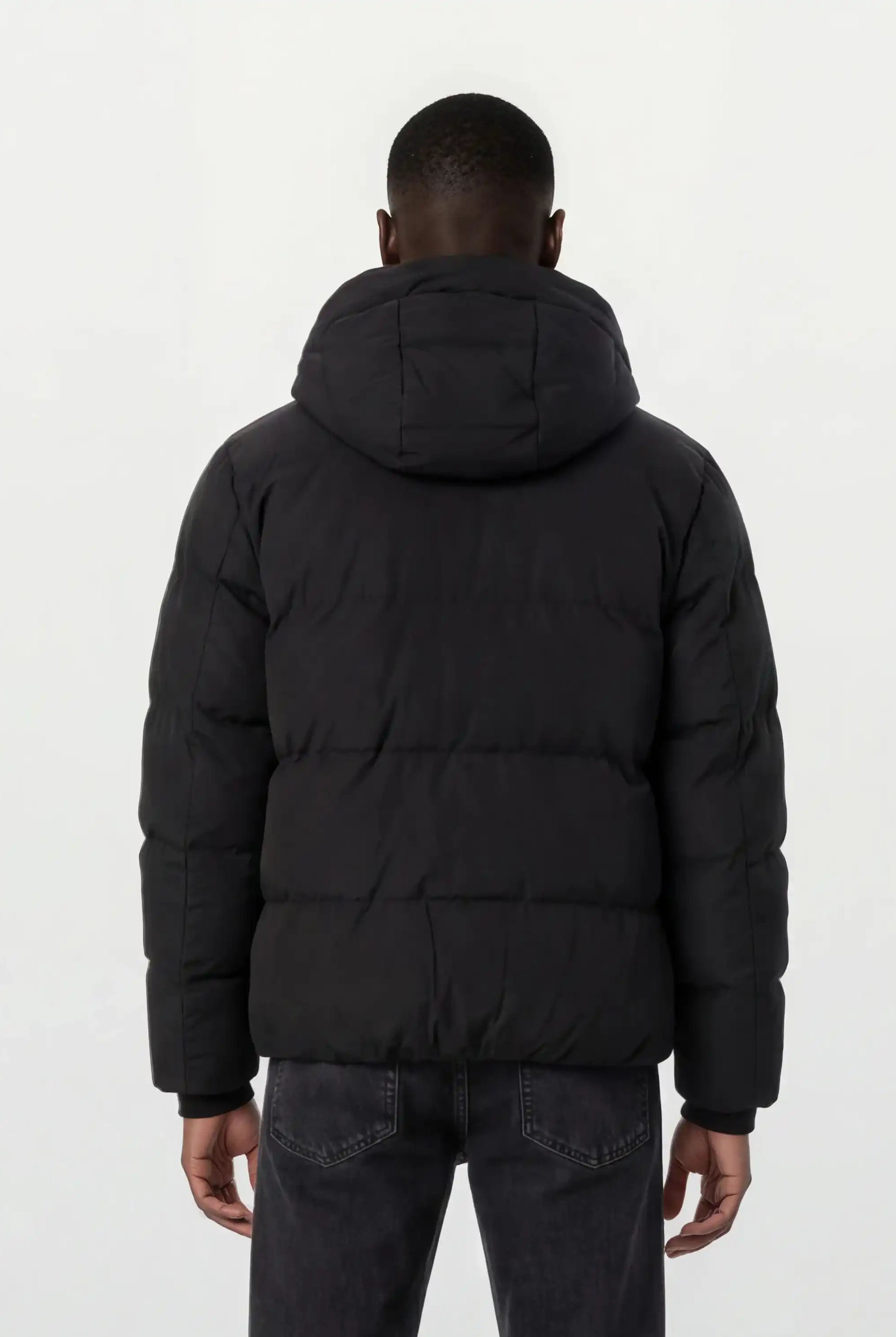 Eine Person mit kurzen Haaren wird von hinten gezeigt, die eine trueprodigy Herren Winterjacke mit fester Kapuze im Oversized Fit und dunkle Jeans trägt. Die Jacke wirkt wasserabweisend, der Hintergrund ist schlicht und hell.