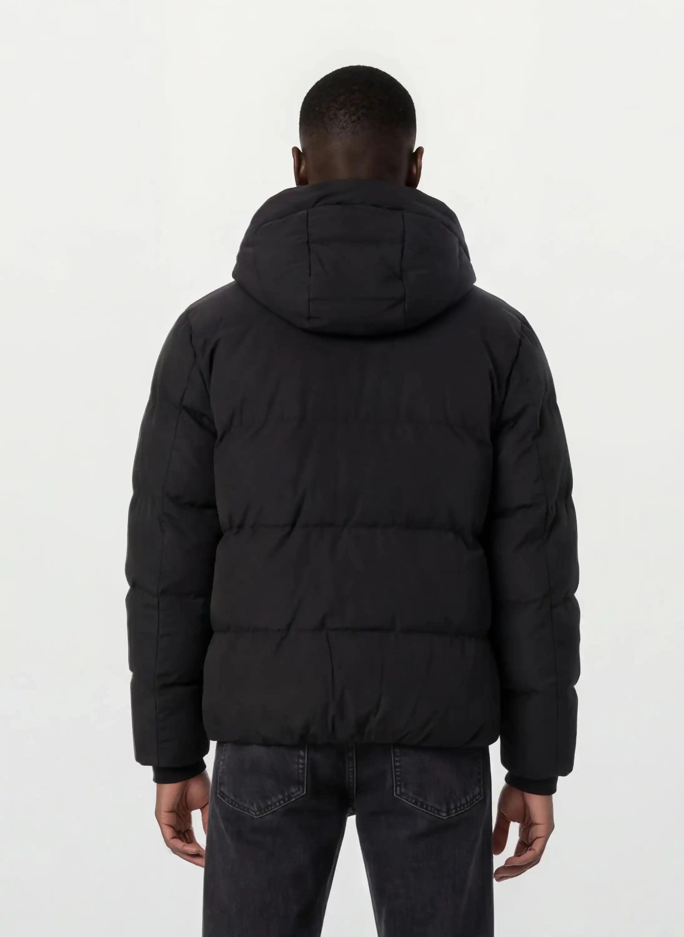 Eine Person mit kurzen Haaren wird von hinten gezeigt, die eine trueprodigy Herren Winterjacke mit fester Kapuze im Oversized Fit und dunkle Jeans trägt. Die Jacke wirkt wasserabweisend, der Hintergrund ist schlicht und hell.