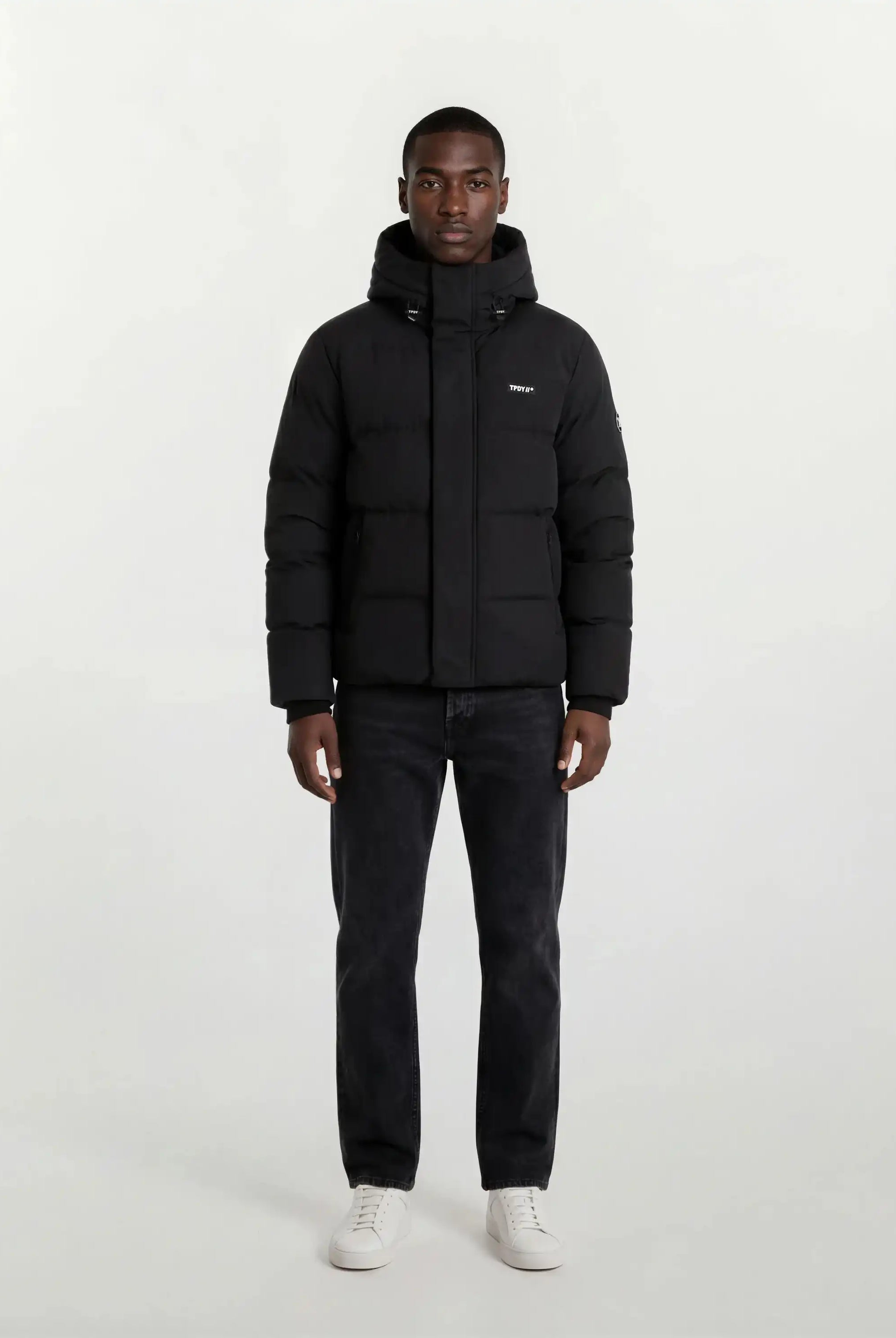 Eine Person steht mit dem Gesicht nach vorne vor einem schlichten weißen Hintergrund und trägt die trueprodigy Herren Winterjacke mit fester Kapuze im Oversized Fit, gepaart mit dunklen Jeans und weißen Sneakers.